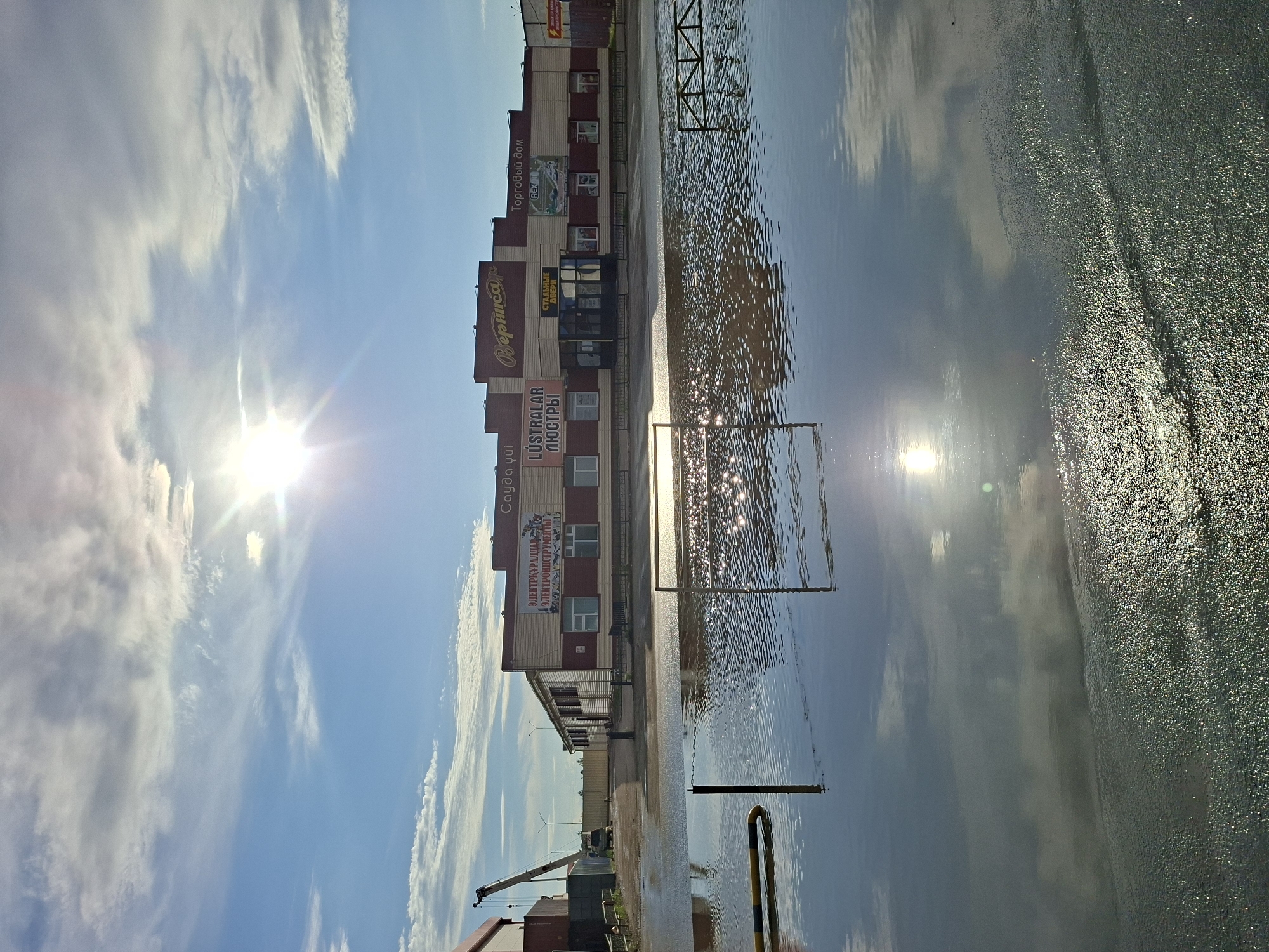 The image depicts a flooded area in front of a commercial building complex. The building has several signs with Russian text, including "Вернисаж" (Vernissage), "Торговый дом" (Trading House), "ЛЮСТРАЛАР ЛЮСТРЫ" (Lustrar Lustry - chandeliers, lamps), and "СТАЛЬНЫЕ ДВЕРИ" (Steel Doors). There are also signs indicating "ЭЛЕКТРОКУРАЛЛАР" (Electrocurallars - possibly electrical goods) and "ЭЛЕКТРОИНСТРУМЕНТЫ" (Electric Tools). The building appears to be a retail establishment. A crane is visible in the background to the left, suggesting construction or industrial activity nearby. The foreground is dominated by a large body of water reflecting the sky and the building. Sunlight is prominently visible in the sky, casting glares on the water's surface. The overall scene suggests a commercial area impacted by flooding. Based on the signage and architectural style, this location is likely in Kokshetau, Kazakhstan.