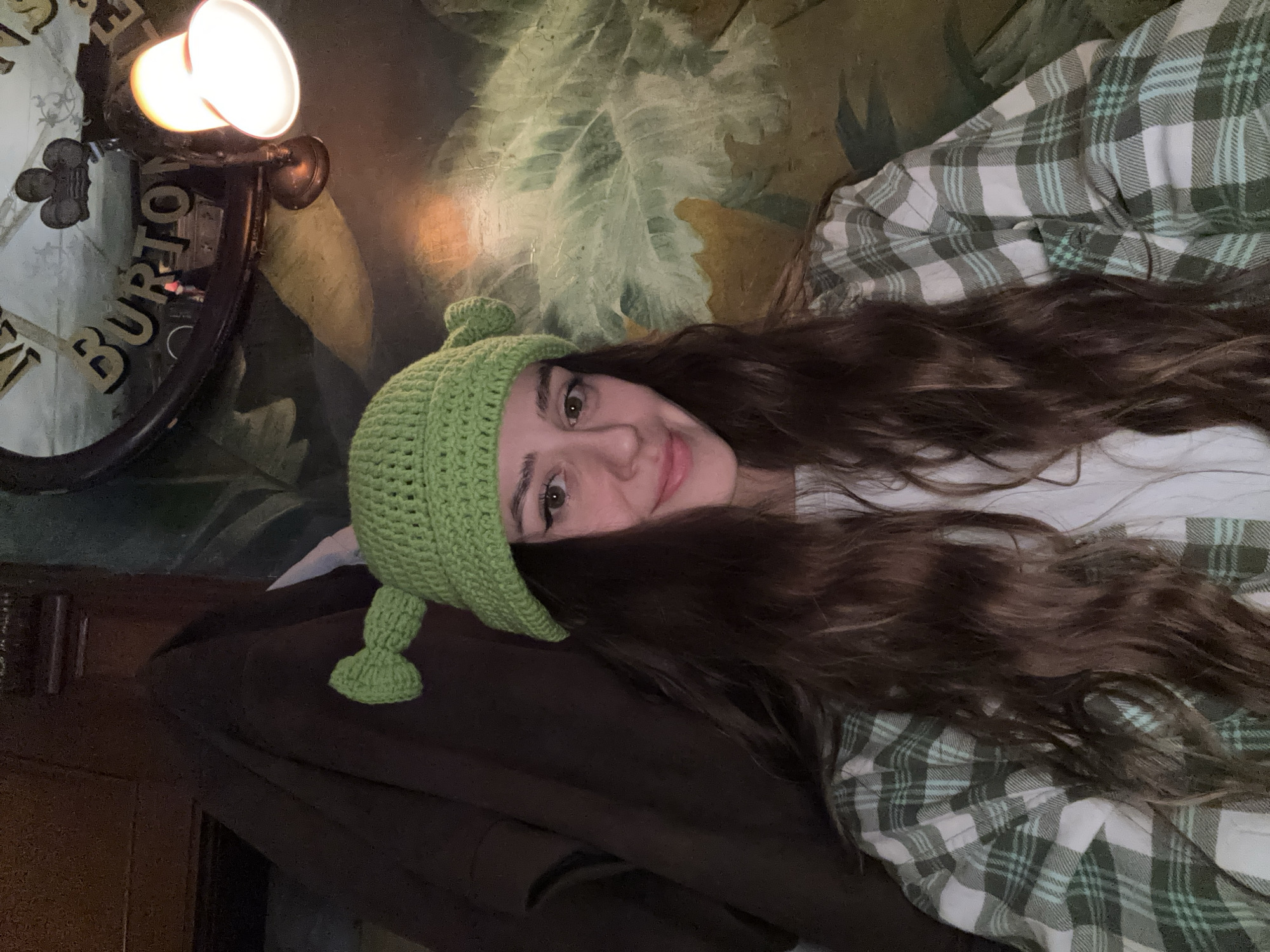 A medium shot depicts a woman lying down. She is wearing a green crocheted hat. The woman has long brown hair, and she is wearing a plaid shirt. Behind her is a wall with a floral pattern and a light fixture. A dark object is visible in the lower left corner of the frame. The image does not show any information about the city of Moscow, Russia.