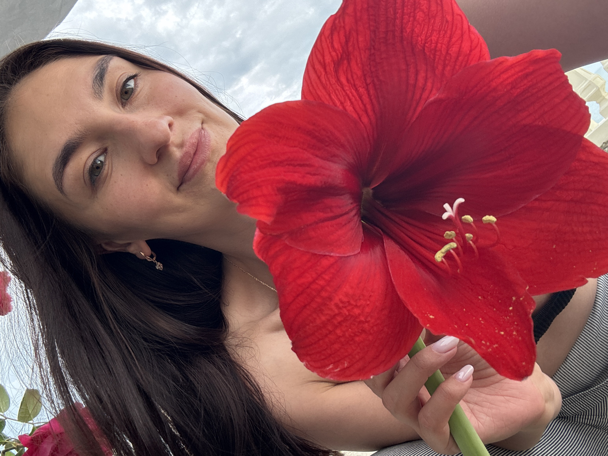 A young woman is photographed in an upward angle close-up shot. She has long dark hair and is wearing small earrings and a delicate necklace. She is holding a large red amaryllis flower with white and yellow stamens close to the camera. The flower dominates the foreground. A striped garment is visible on her body. The background shows a cloudy sky. The location, Sochi, Russia, is not discernible from the image.