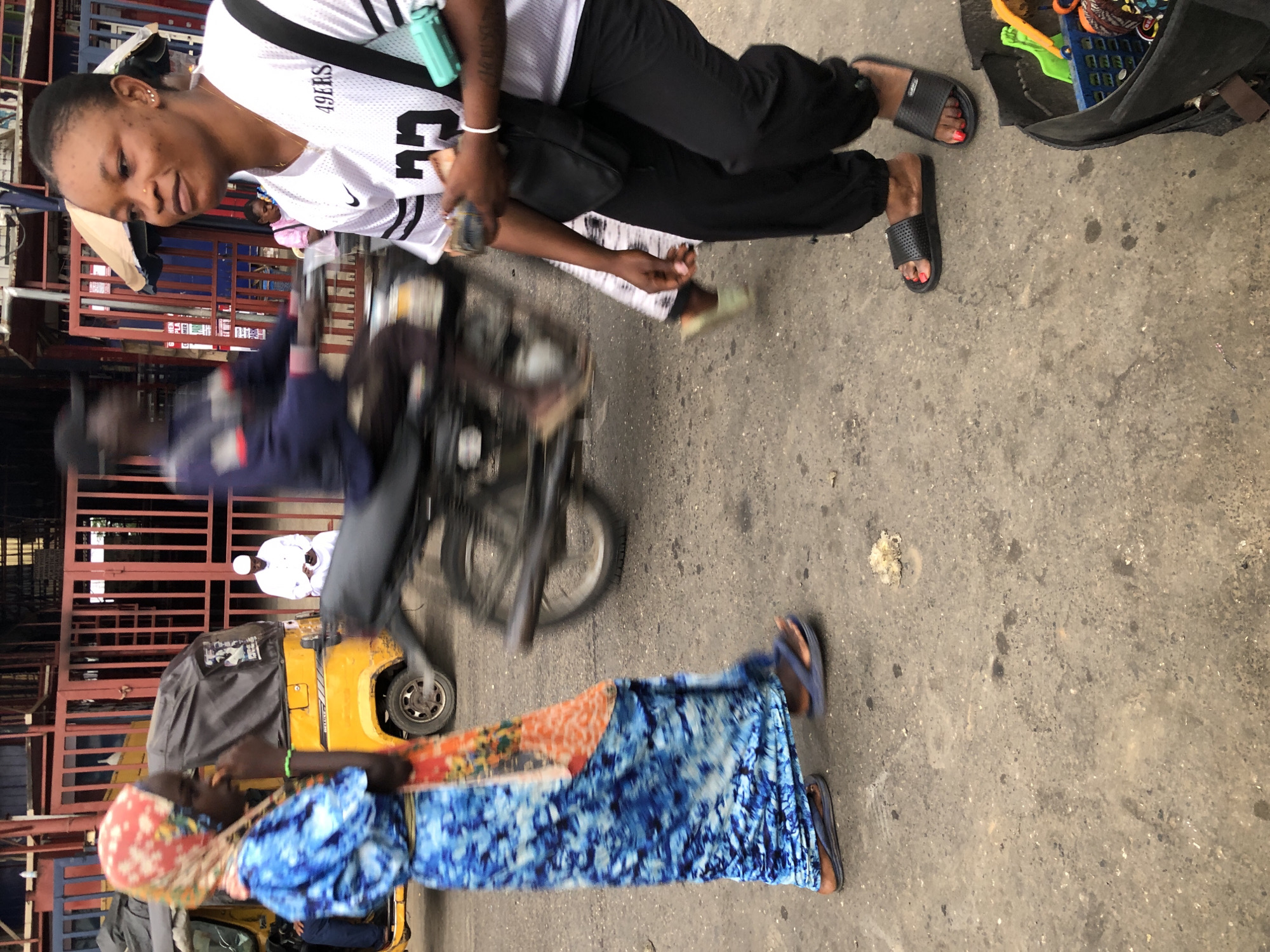 This image captures a street scene in Ikeja, Nigeria. In the foreground on the right, a woman wearing black slide sandals with red nail polish is standing. She has a black crossbody bag slung over her shoulder and is holding some cash. Her shirt displays "49ERS" and the number "23". To her left, a person wearing a blue and white tie-dye dress and a red headscarf walks away from the camera, wearing blue flip-flops. Behind them, a yellow auto-rickshaw (Keke Napep) is parked. In the middle ground, a person is riding a motorcycle, their face blurred by motion. Further in the background, a person in white Islamic attire is seated and looking at a mobile phone. The environment appears to be a busy urban street with various structures and red metal gates visible. The ground is paved with asphalt.