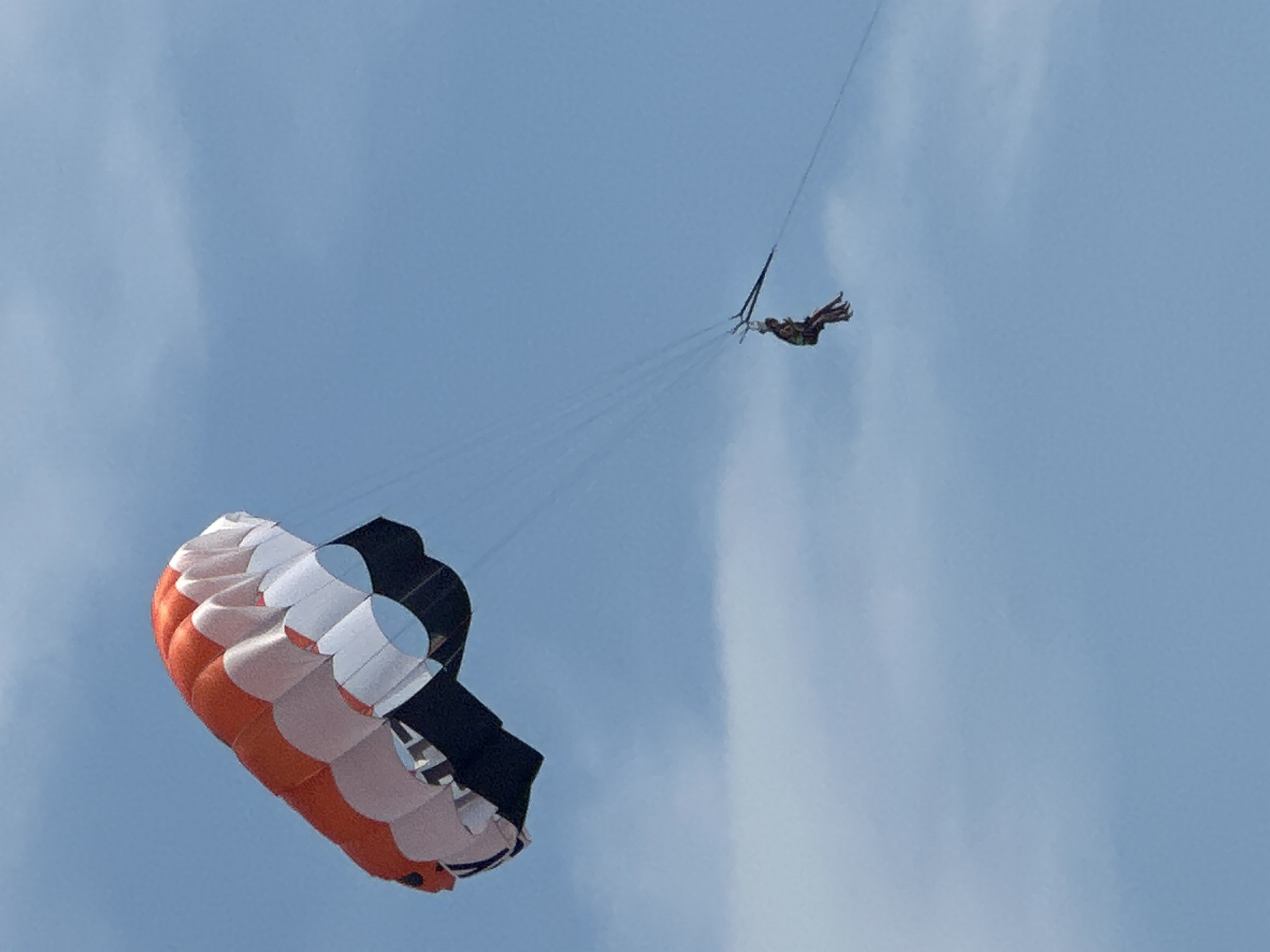 In the sky above Boreti, Montenegro, a parasail with orange, white, and black panels is inflated and visible. Below the parasail, two individuals are seated, suspended by tethers. The sky is a pale blue with wispy white clouds, indicating daylight conditions. The perspective suggests the photo was taken from below, looking upwards.