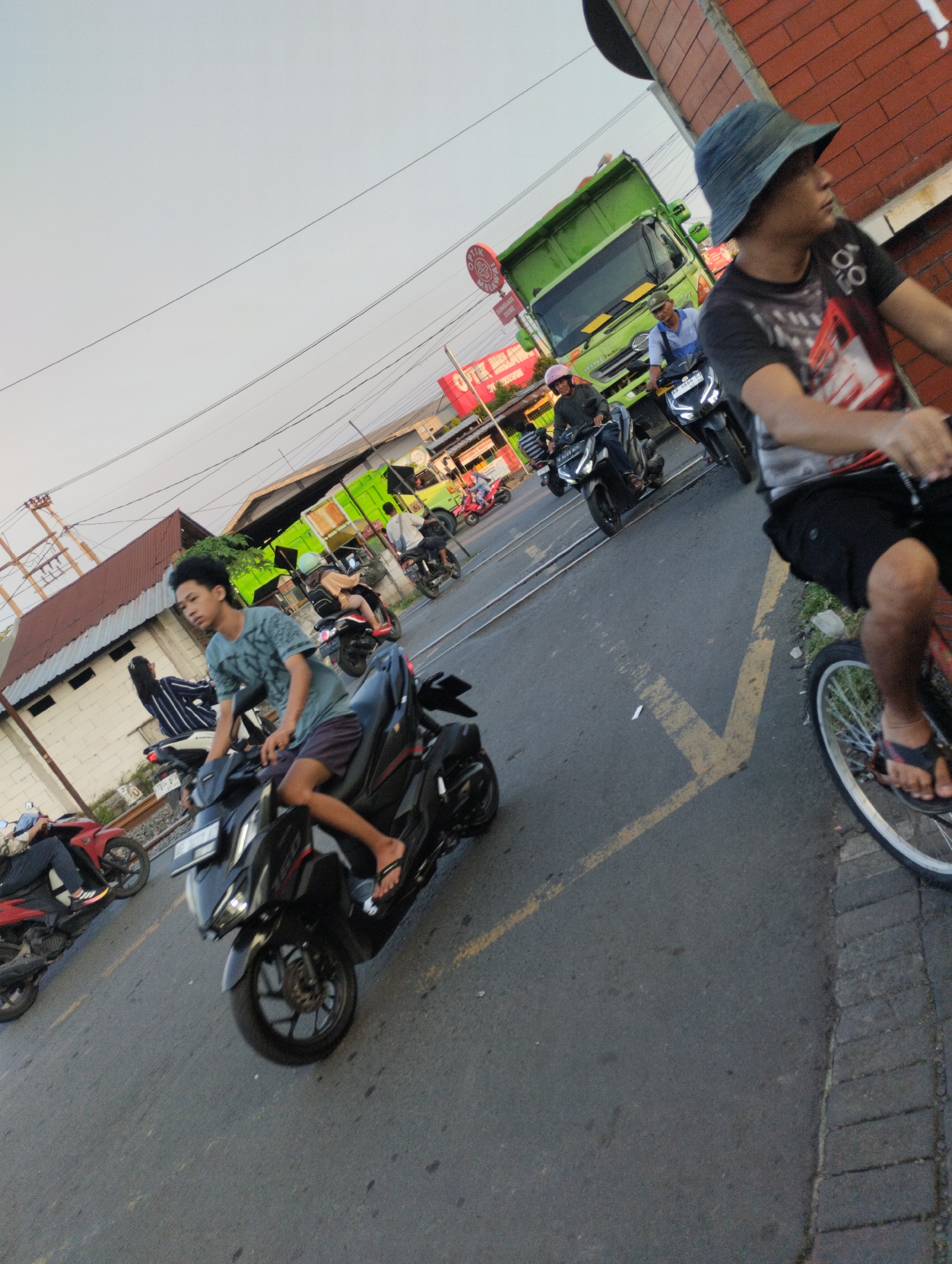 The image depicts a street scene in Bringinbendo, Indonesia, likely during the late afternoon or early evening, given the soft, warm lighting. The setting appears to be a moderately busy road with several vehicles and pedestrians. The foreground features a young man riding a black scooter. He is wearing a blue-green patterned t-shirt and dark shorts, with flip-flops on his feet. Behind him, further down the road, is a bright green truck, its engine compartment open. To the right of the truck, another person is seen on a scooter, wearing a helmet.
To the far right of the frame, a person is riding a bicycle, seen from the chest down. They are wearing a dark t-shirt and black shorts, with sandals. The bicycle has a silver rim and spokes. In the background, there are buildings with red tile roofs and various shops or stalls lining the street, one of which has a red circular sign with white text, likely a business logo. Power lines are visible crossing the sky.
Several other individuals are present on scooters and motorbikes, indicating a typical urban or semi-urban traffic environment. One scooter in the middle ground has a rider with a pink helmet, and another further back has a rider in a light-colored shirt. A red scooter is parked or moving on the left side of the image. The road surface is asphalt, with yellow lane markings. The overall atmosphere suggests a normal day with ongoing traffic and daily life activities.