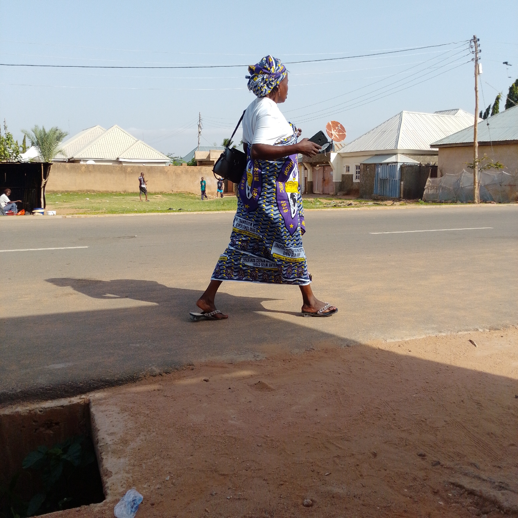 In Gombe, Nigeria, a woman walks along a paved road, her body angled towards the right of the frame. She wears a white shirt and a vibrantly patterned wrap skirt in shades of blue and yellow, with a matching headscarf. A black bag is slung over her left shoulder, and she holds a dark-colored smartphone in her right hand. Her footwear consists of patterned flip-flops. In the background, several buildings with light-colored metal roofs are visible, interspersed with a dirt wall and vegetation. A wooden pole with electrical wires stands on the right side of the image. The ground in the foreground is a reddish-brown dirt path. A man sits in the shade of a structure to the left, and other figures are discernible further in the distance. The sky is clear and blue.