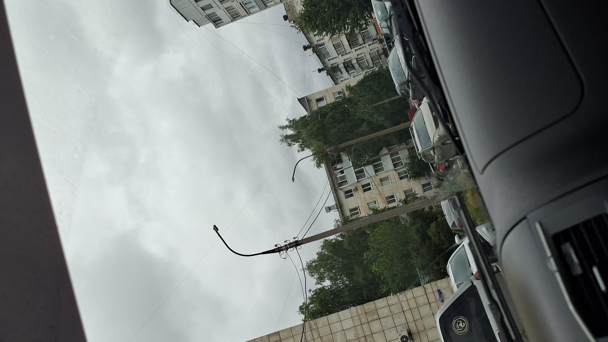The image depicts a cloudy sky over a city street in Chaikovsky, Russia. In the foreground, the interior of a vehicle is visible, with part of the dashboard and windshield. Through the windshield, a row of apartment buildings and trees can be seen. Two lampposts with curved tops stand prominently on the street, with power lines running between them. Several cars are parked or driving along the street, including a light-colored SUV and darker sedans. The buildings appear to be multi-story residential structures, typical of urban areas. The overall atmosphere is overcast and somewhat muted.