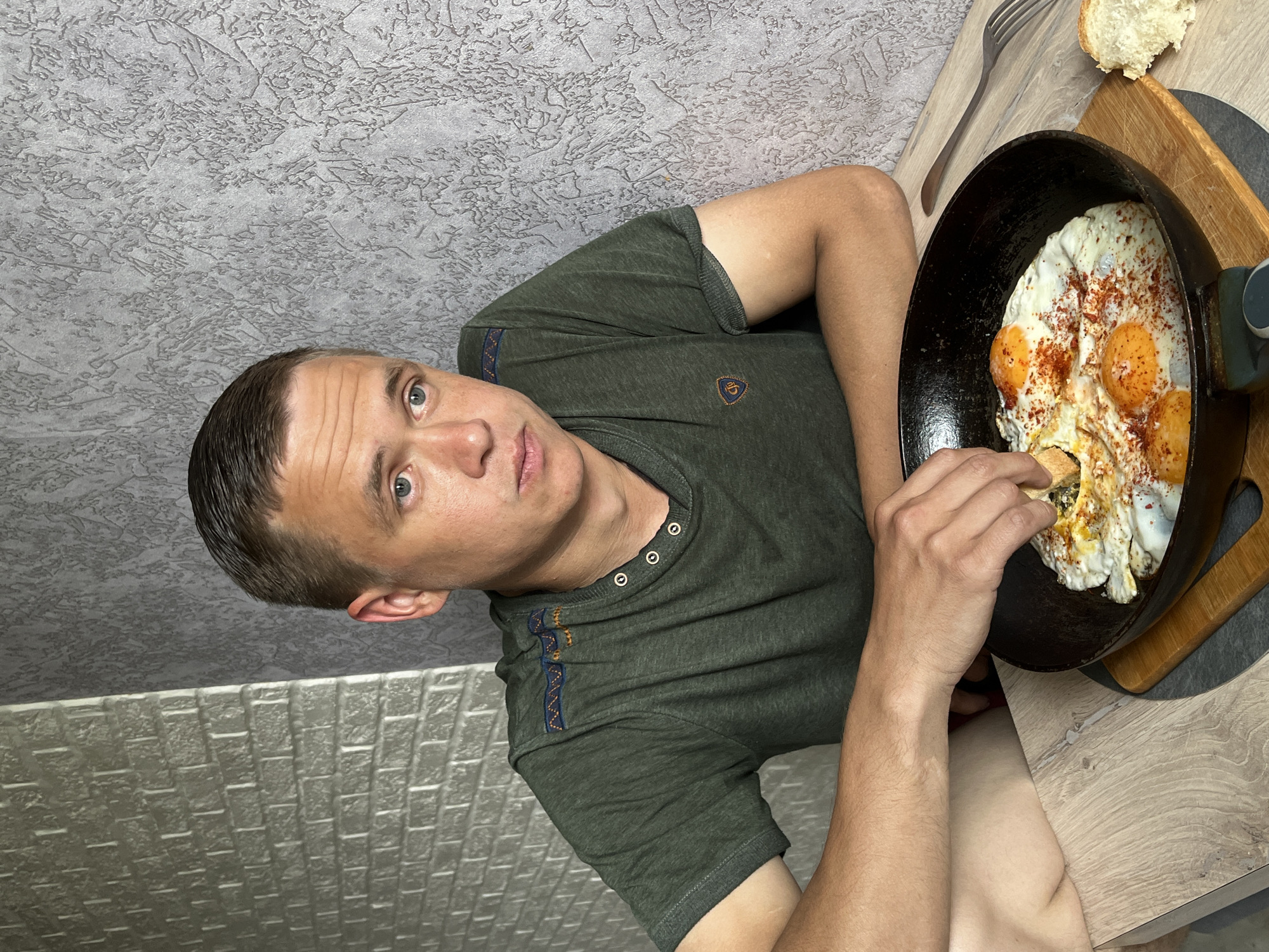 A man is seated at a table in what appears to be a residential interior, likely in the city of Chaikovsky, Russia given the query context, although this cannot be definitively confirmed from the image alone. He is wearing a dark green t-shirt with a buttoned placket. In front of him, a cast iron skillet rests on a wooden cutting board. The skillet contains fried eggs, sprinkled with a red spice. The man is holding a piece of bread in his right hand, seemingly about to dip it into the eggs. A fork and a piece of bread are also present on the table near the skillet. The wall behind the man is textured and grey. The wall to his left has a brick-like pattern. The table surface is a light wood grain.