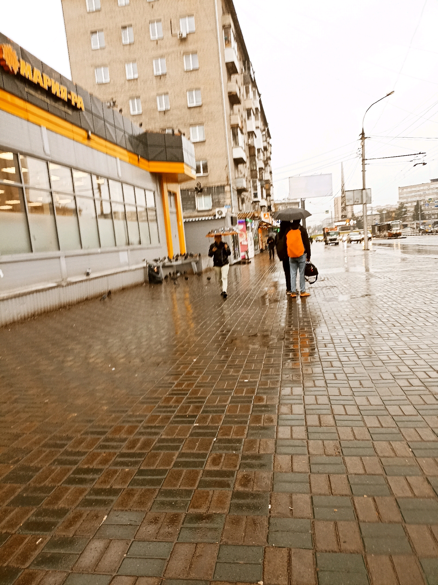 The original image is an eye-level, medium shot taken on a cloudy day in an urban environment. The foreground is dominated by a wet brick sidewalk with a pattern of alternating brown and dark gray bricks. In the background, a modern building with a gray facade and a prominent yellow and orange architectural detail extends across the frame. Above the building, a grey sky is visible.
To the right of the frame, two people are walking away from the camera, one holding a black umbrella over both of them. The person closer to the camera is wearing dark pants and a bright orange backpack. The person behind them is wearing dark pants. To the left of these individuals, another person is walking towards the camera, holding a translucent umbrella and wearing a dark jacket and white pants.
Further in the background, to the right, a street with cars and a bus is visible, with utility poles and wires overhead. The overall impression is of a city street on a rainy day.