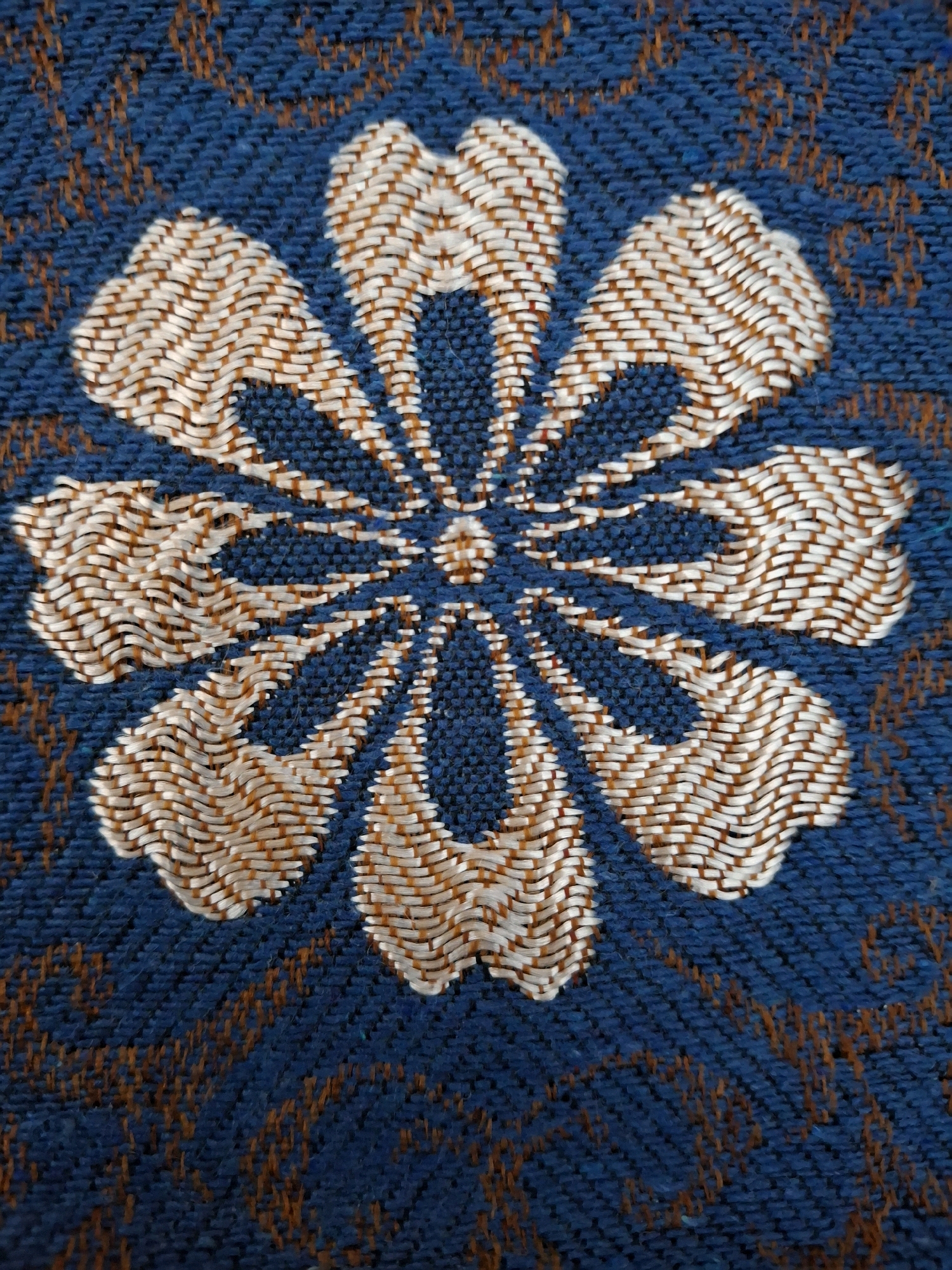 The image is a close-up, high-angle shot of a blue textile with an intricately woven floral pattern. The flower, rendered in white and brown threads, has eight petals radiating from a central point. The petals are detailed with a woven texture that creates a sense of depth and dimension. The background is a solid blue fabric with a subtle, darker patterned texture that appears to be woven into the material itself, possibly a damask or jacquard weave. The threads are visible, showing the texture of the fabric. There are no people, animals, or any objects other than the textile visible in the image. The lighting is even, highlighting the textures and patterns without harsh shadows. There is no information in the image that allows for a connection to Lumajang, Indonesia.