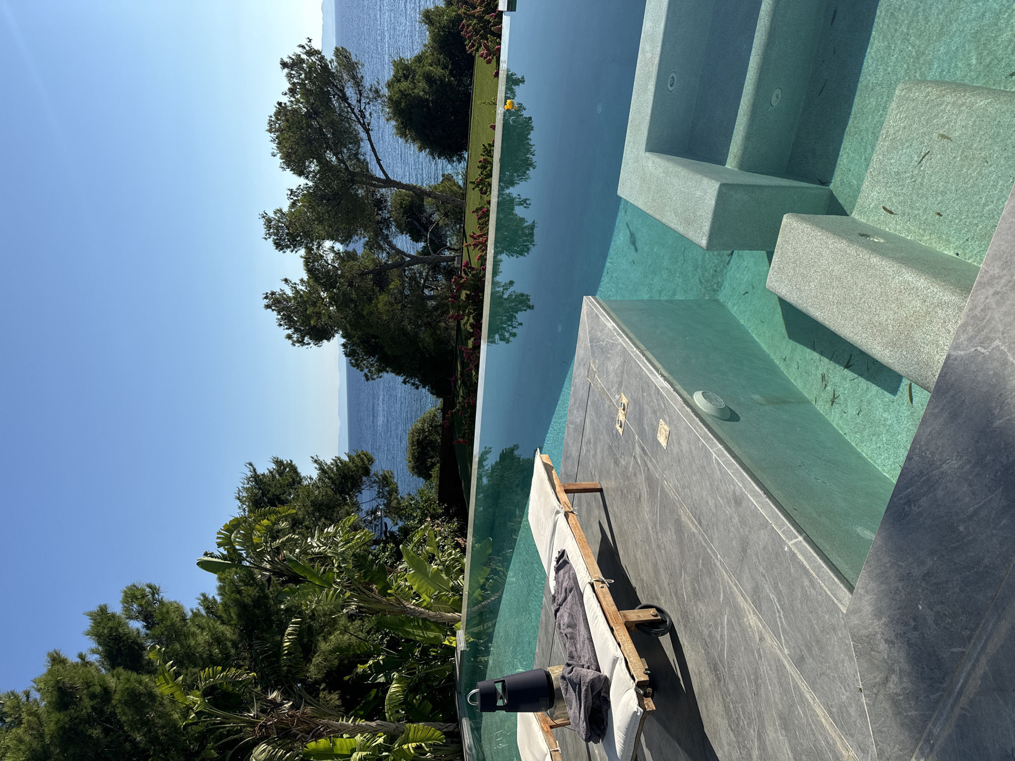 The image depicts a luxurious infinity pool overlooking the Aegean Sea, likely situated in the Vouliagmeni Municipal Unit of Greece. The pool is a prominent feature, with its turquoise water reflecting the clear blue sky and lush green trees. In the foreground, a modern lounge chair with a white cushion and folded grey towels sits on a grey stone deck. A black portable speaker is placed on the lounge chair. The pool's edge is lined with stone, and steps descend into the shallower end of the pool, where small fish are visible. Beyond the pool, a well-maintained lawn is bordered by vibrant red flowering bushes and tall pine trees. The sea stretches out to the horizon, with distant landforms visible under a cloudless sky. The scene exudes tranquility and opulence, suggesting a high-end resort or private residence.