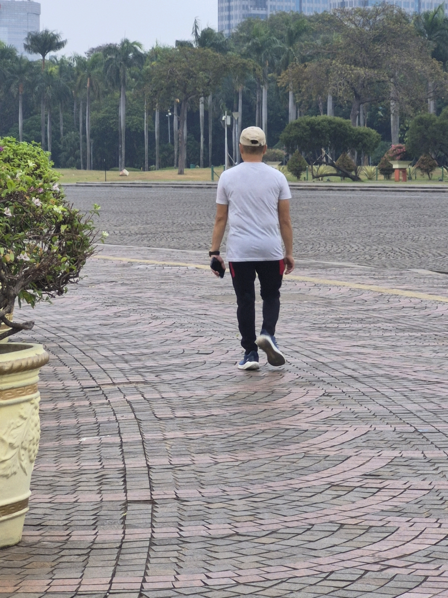 A man wearing a white t-shirt, black pants with red accents, a beige cap, and blue sneakers is walking away from the camera on a patterned brick walkway. He is holding a black smartphone in his right hand. In the background, there is lush green foliage with tall palm trees, suggesting an outdoor park or garden setting. The sky is overcast. The scene is located in Central Jakarta, Indonesia, in an area that appears to be a public park or plaza with decorative planters.