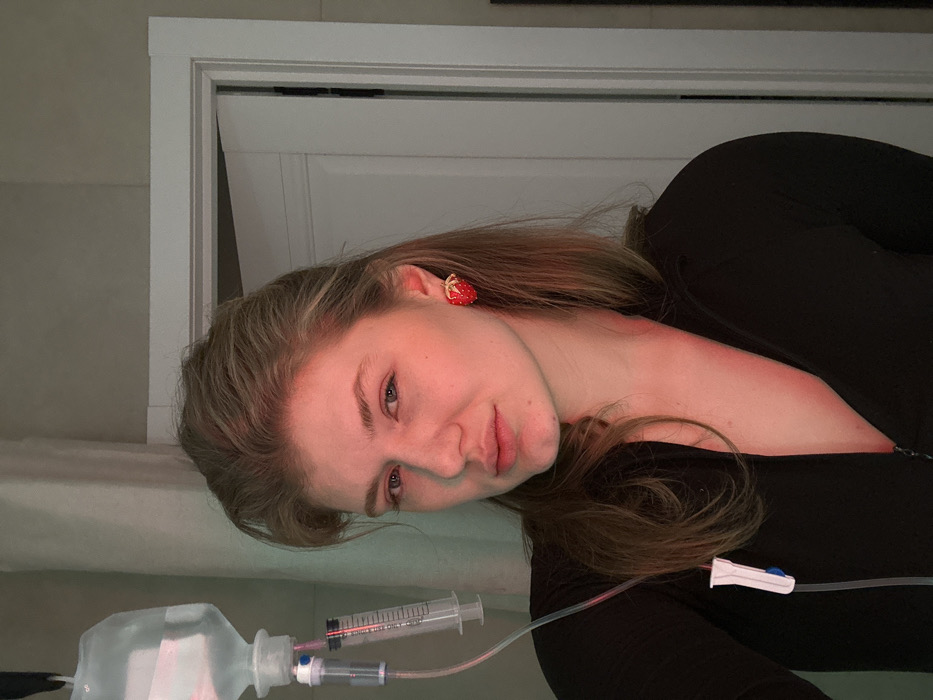 A female individual is depicted indoors, in what is identified as Kazan, Russia. The subject, an adult with light brown hair, is wearing a dark, possibly black, zip-front top. A single red strawberry-shaped earring is visible on her right ear. Her gaze is directed towards the camera, maintaining a neutral expression. An intravenous (IV) fluid bag containing a clear liquid is suspended in the upper left quadrant of the frame. An IV line descends from the bag, incorporating a clear syringe with measurement markings and a white plastic clamp with a blue roller, suggesting ongoing fluid administration. The specific point of venous access is not visible. The background consists of light-colored walls and a partially open white door frame. Illumination creates a prominent red hue across the subject's face and upper body, with a faint greenish cast evident in the background.