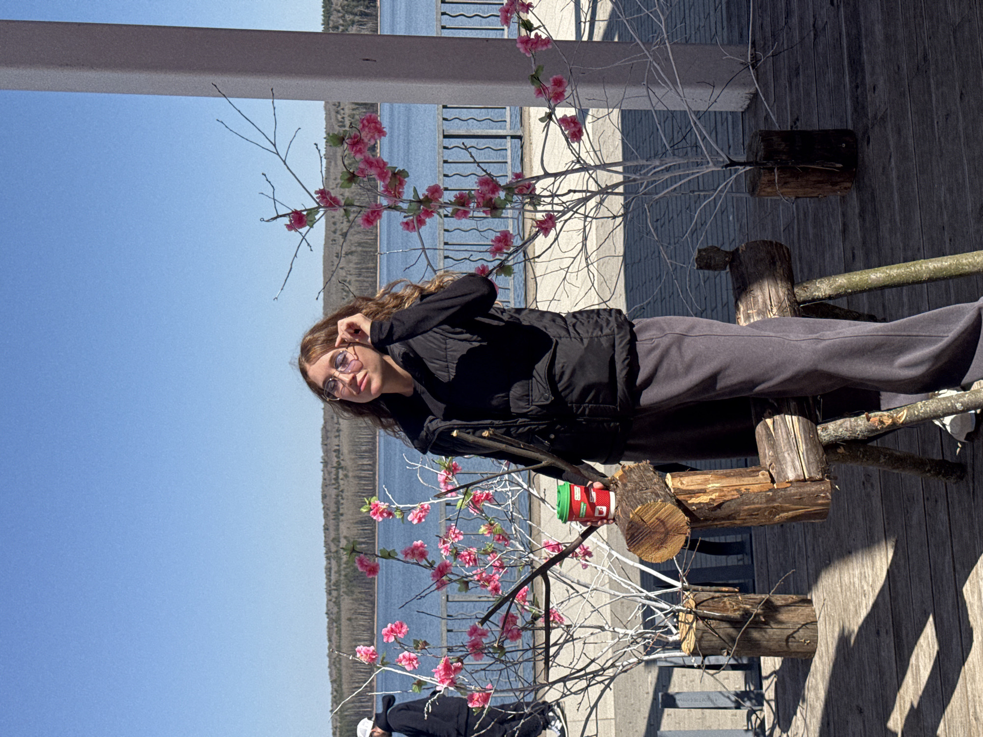 The image depicts a young woman in Chaikovsky, Russia, posing near an artificial cherry blossom tree on a wooden boardwalk. The woman is wearing glasses, a black jacket, and dark pants. She appears to be holding up her glasses. In the background, a body of water and treed land are visible under a clear blue sky. A coffee cup is on the tree. The boardwalk is constructed of wooden planks. The decorative tree is planted in a log planter. Other people are partially seen in the foreground.