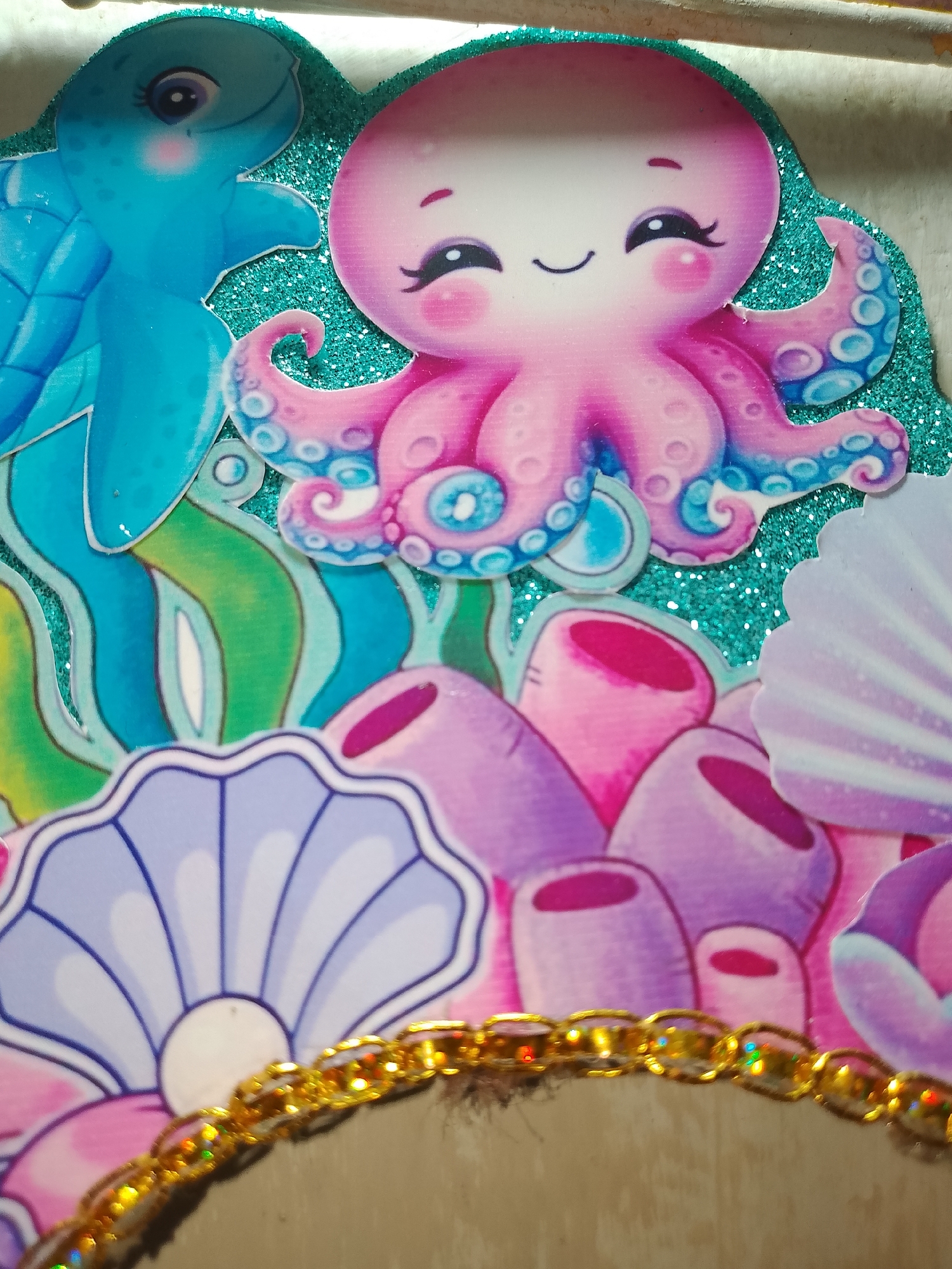 The image presents a detailed close-up of a multi-layered decorative item with an aquatic theme, located in Santa Ana, Venezuela. The central focus is an anthropomorphic pink octopus with blue tentacle details, featuring a stylized smiling face. To the immediate left and partially obscured is a light blue sea turtle, also depicted with a friendly expression. Both figures are cut-out elements layered over a background. The background consists of a shimmering, teal-colored glitter application, simulating water. Below the main animal figures, additional marine elements are visible, including stylized green and blue seaweed patterns, several pink and purple coral formations, and multiple fan-shaped seashells in shades of purple and white. A golden, iridescent chain forms a horizontal border across the lower section of the image, acting as a foreground element. The composition suggests a crafted or designed display.