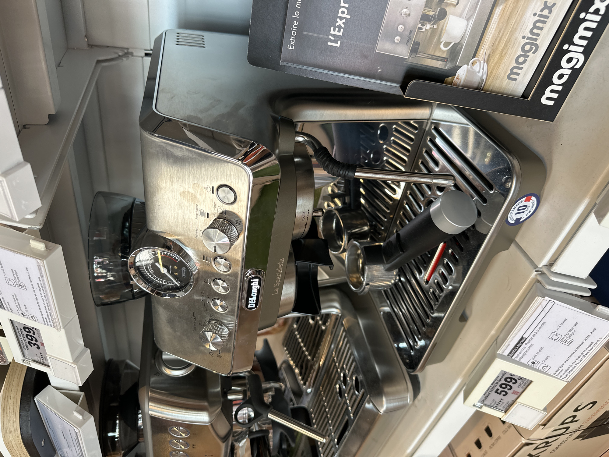 This media file is a close-up shot of a silver DeLonghi La Specialista espresso machine displayed on a shelf, likely in a retail setting. The machine has a sleek, modern design with prominent stainless steel elements, including the portafilter, drip tray, and control panel. The control panel features several buttons, a dial with settings like "Espresso," "Americano," and "Hot Water," and a prominent pressure gauge. A steam wand is attached to the side of the machine.
Next to the espresso machine, there are product information tags and boxes. One tag displays a price of "399" and another shows "599.99". Another visible tag is for a "Magimix" product, featuring images of coffee cups. There are also boxes with branding that includes "KRUPS." The overall scene suggests a product display in a store, possibly an electronics or home appliance store. The lighting appears to be typical indoor retail lighting. There is no indication of people or a specific time of day or weather conditions. The focus is entirely on the product and its associated marketing materials.