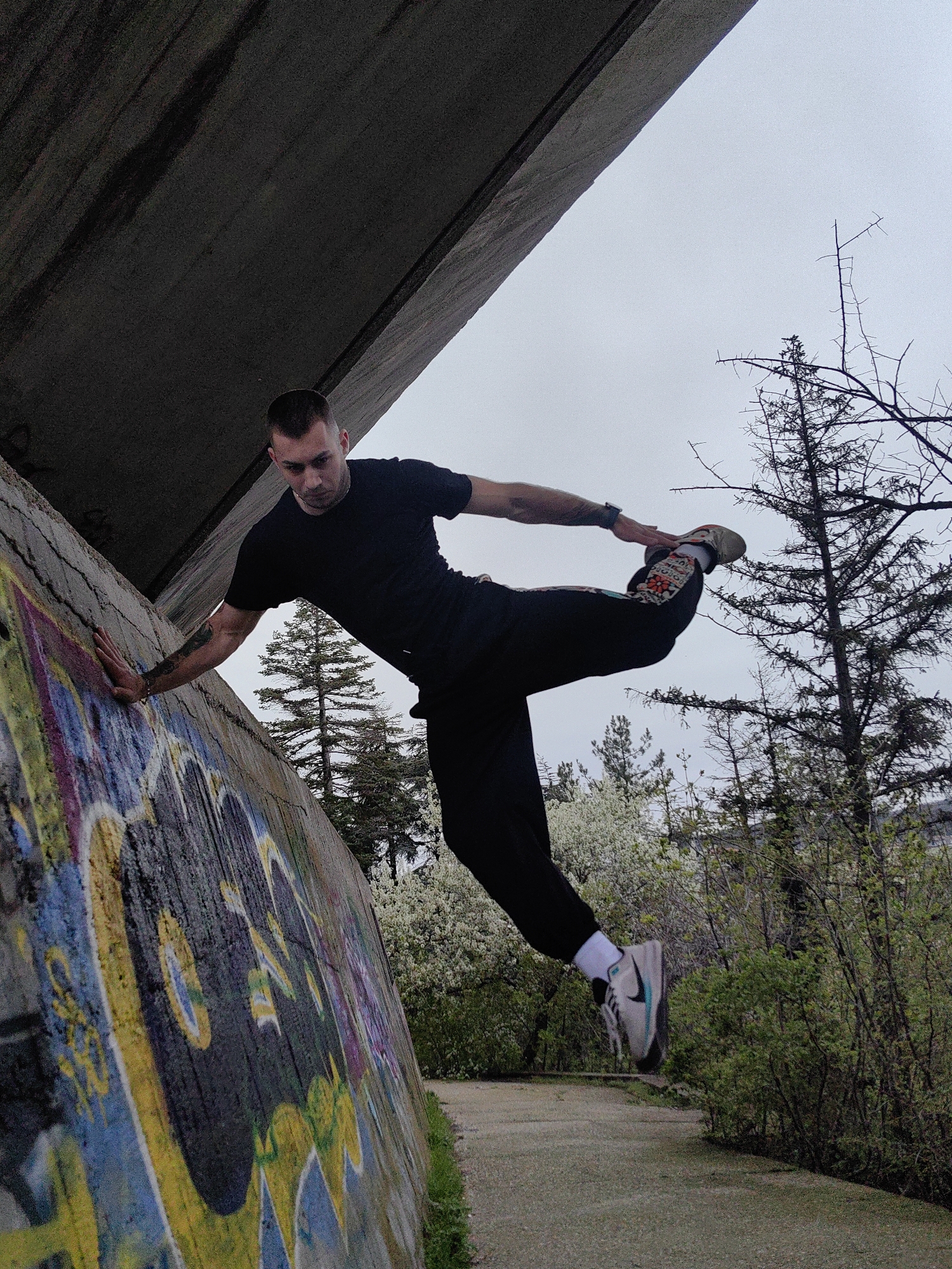 A man is captured mid-motion in an acrobatic pose. He wears a black t-shirt, black pants with decorative trim on one leg, white socks, and sneakers. His left hand is pressed against a concrete wall covered in graffiti. His right leg is extended behind him, with his right hand touching his foot. The background reveals trees and foliage, suggesting an outdoor park or urban environment. A concrete overhang or bridge structure frames the scene from above.
The setting is likely within an urban park landscape in Varna, Bulgaria. Graffiti on the wall suggests an urban setting.