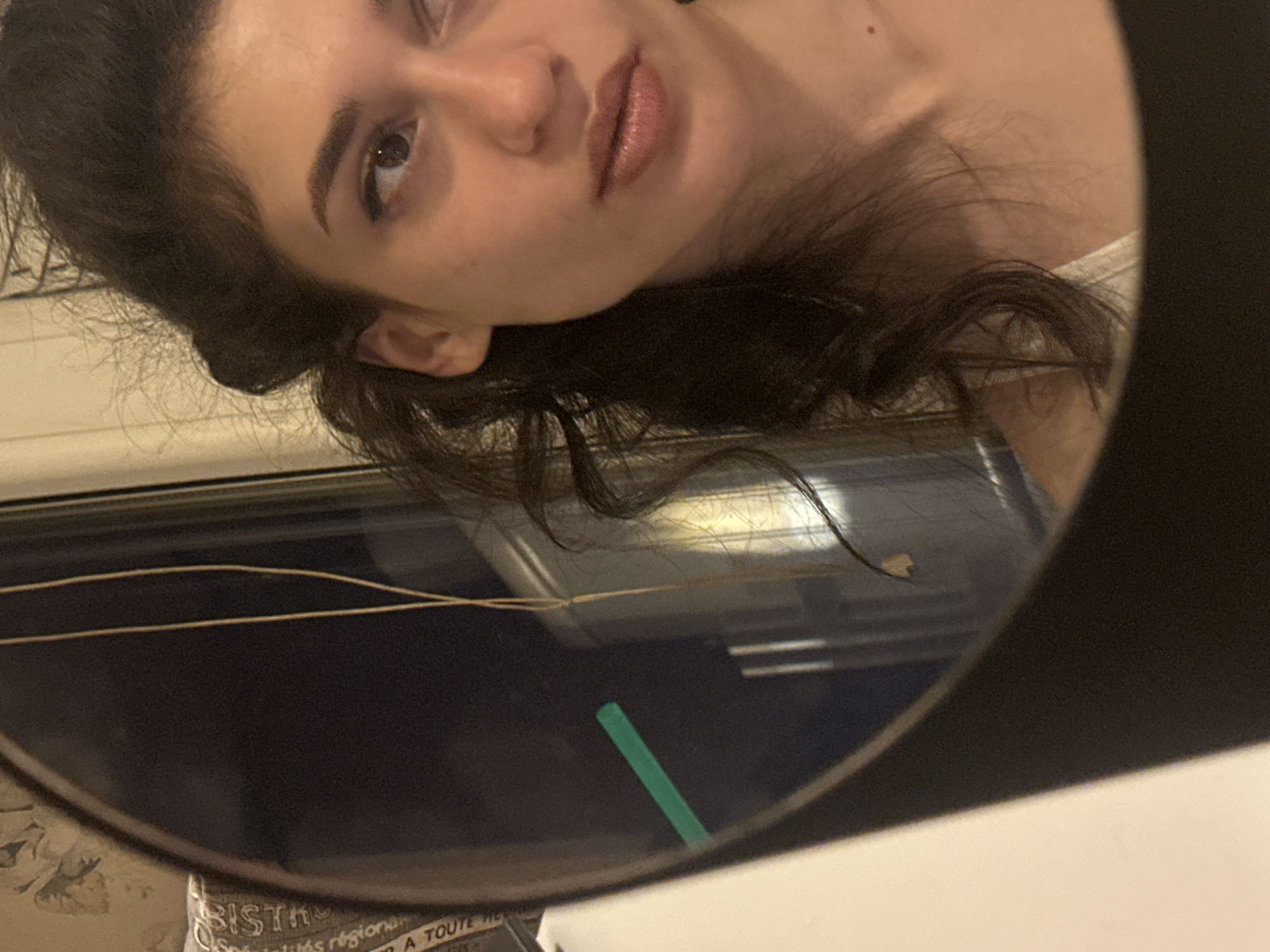 The image shows a young woman with dark hair, looking into a mirror. She is wearing a white top. Her reflection is visible in the mirror, and part of her face and hair are also captured outside the mirror. There is a green straw visible in the mirror's reflection. The background appears to be indoors, with some faint writing visible on a surface in the reflection. The image does not contain any information about the city of Sofia, Bulgaria.