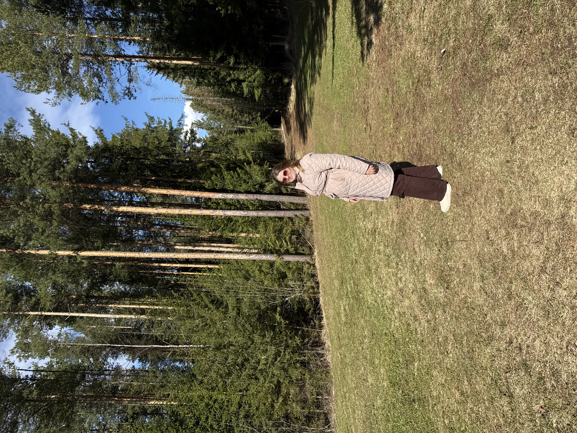 A landscape photograph shows a person standing in a grassy field next to a dense forest. The person is wearing sunglasses, a light beige quilted jacket, dark brown pants, and light-colored shoes. The forest to the left is composed of tall evergreen trees, with glimpses of blue sky visible between the branches. The grassy field has patches of green and brown, suggesting a transition between seasons. The image appears to be taken in daylight, with shadows indicating the direction of the sun. The location is in the city of Адам, Russia.