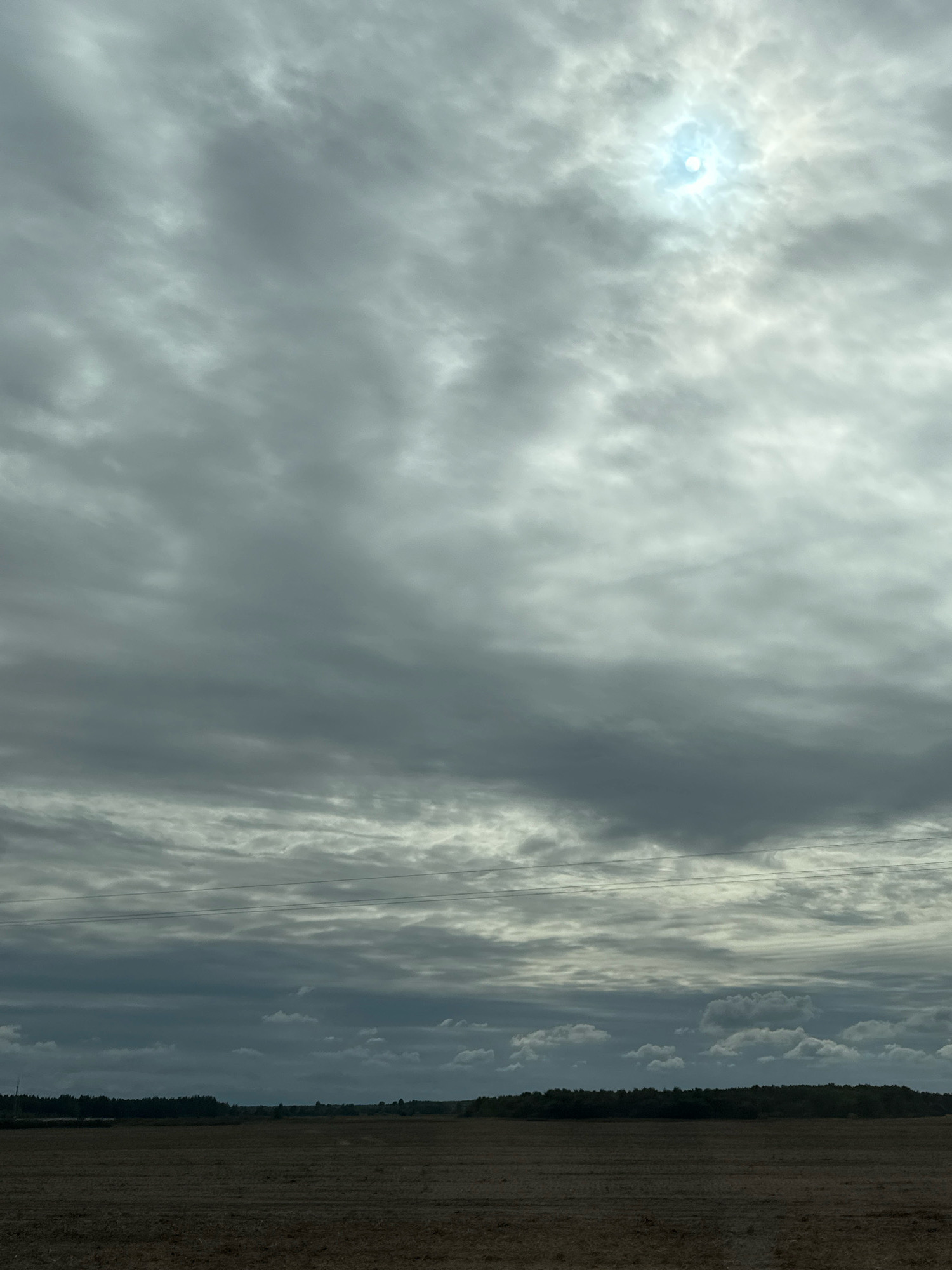 The image depicts a rural landscape under a cloudy sky with the sun partially visible. The foreground is a dry, harvested field, likely agricultural. In the mid-ground, a dark line of trees forms the horizon, with a few sparse structures and possibly a small body of water visible to the left. Overhead, the sky is filled with a thick layer of grey clouds, with lighter patches indicating where the sun is breaking through, creating a diffuse, bright spot. Two thin, dark horizontal lines, likely power lines, stretch across the sky in the middle distance. The overall scene is overcast and somber, characteristic of a cloudy day in a rural setting in Краснаўскі сельскі Савет, Belarus. There are no people or animals visible in the image.