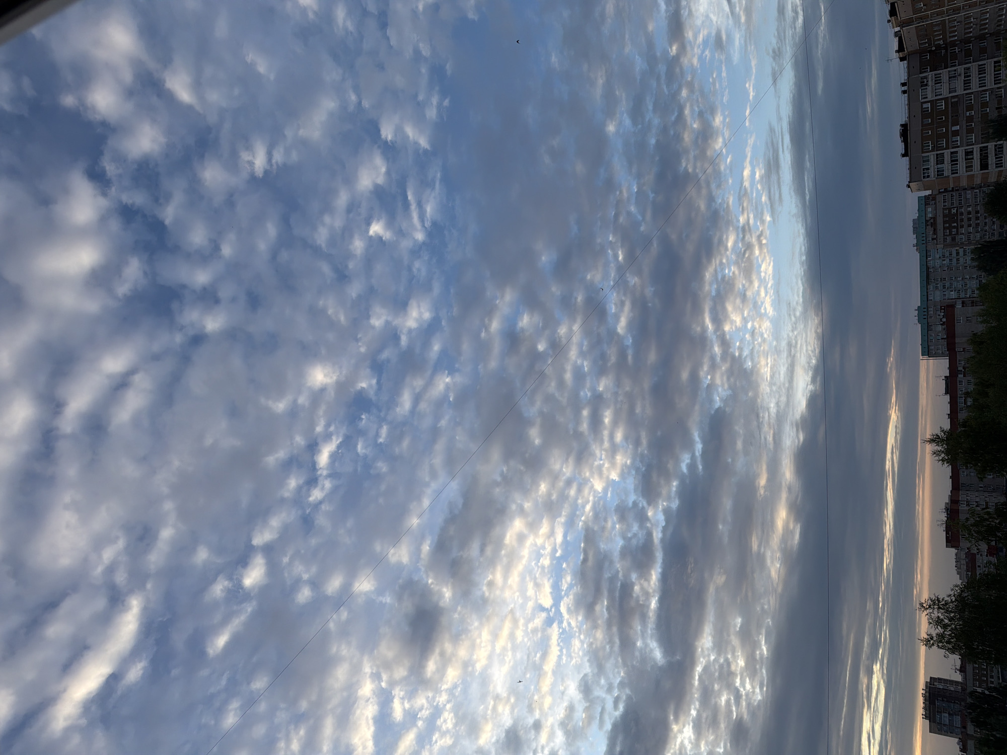 A medium shot captures a wide view of the sky above Izhevsk, Russia, during what appears to be a sunrise or sunset. The sky is filled with scattered clouds, varying in density and illumination. Some clouds are bright white, catching the light, while others are tinged with gray and blue. A thin, dark wire stretches diagonally across the upper portion of the frame, from the top left to the middle right.
Below the sky, the upper portions of several multi-story buildings are visible, suggesting an urban environment. These buildings have a mix of architectural styles and colors, with some featuring lighter facades and others darker. Trees are also present, with their tops reaching up towards the lower edges of the buildings. The overall scene is one of a city skyline beneath a dynamic sky.