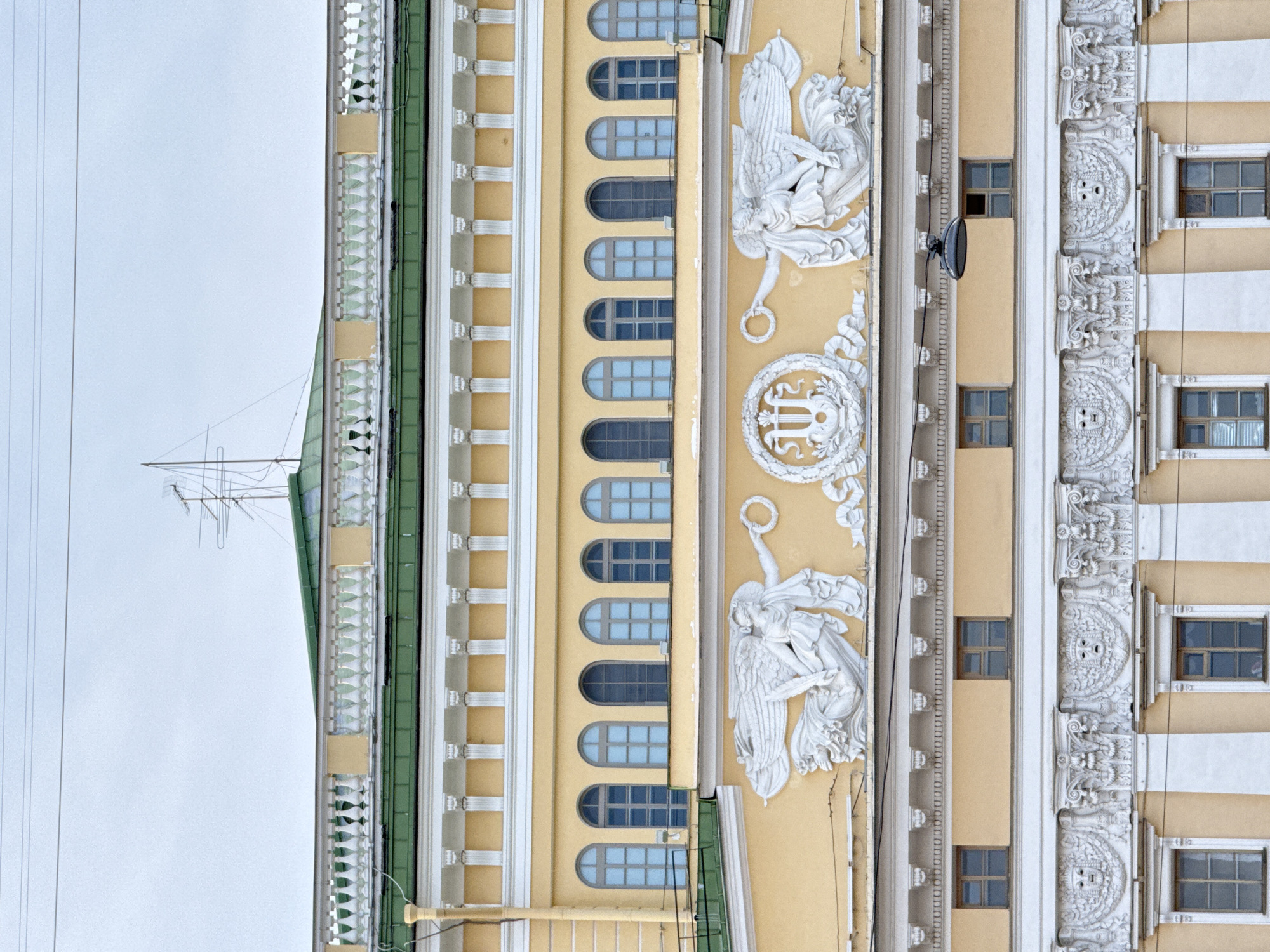 The image depicts a partial view of the facade of a building in Saint Petersburg, Russia. The building exhibits ornate architectural details, including decorative moldings, sculpted figures, and rows of arched windows. The color palette consists primarily of pale yellow, white, and green. The sky is a muted grey, suggesting overcast conditions. An antenna is visible atop the structure. There is no indication of human presence within the frame.