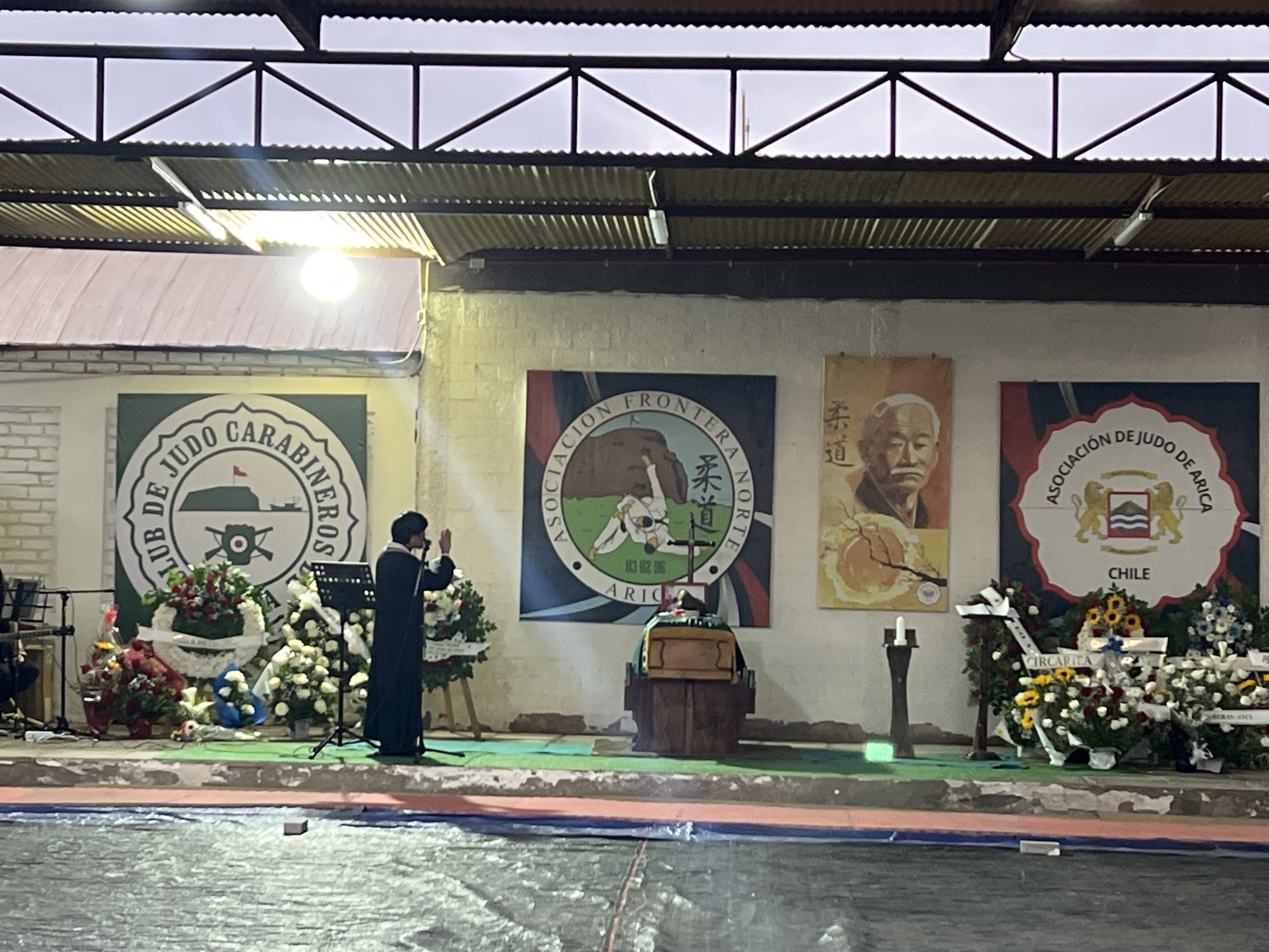 The image depicts an outdoor ceremony under a corrugated metal roof structure, likely in Arica, Chile, given the banners present. In the foreground, a person dressed in a dark robe stands facing a wooden coffin adorned with a green cloth. The individual is gesturing with their right hand, appearing to be speaking or leading a prayer. Behind the coffin, a lit candle stands next to a tall, dark fixture. The scene is surrounded by numerous floral arrangements, including wreaths and bouquets of white, red, and yellow flowers, some bearing white ribbons with text.
To the left, a banner with a circular design features text in Spanish and an emblem that includes a tank. Further to the left, a musician can be seen with instruments. To the right of the coffin, another banner displays a crest with two lions and the word "CHILE" below, along with text that appears to be "ASOCIACIÓN DE JUDO DE ARICA". A portrait of an elderly Asian man is visible on a banner to the right of the central judo association banner. The ground is covered with what appears to be a blue and red tarp in the immediate foreground, and green mats further back. The overall atmosphere suggests a solemn occasion, possibly a memorial service.