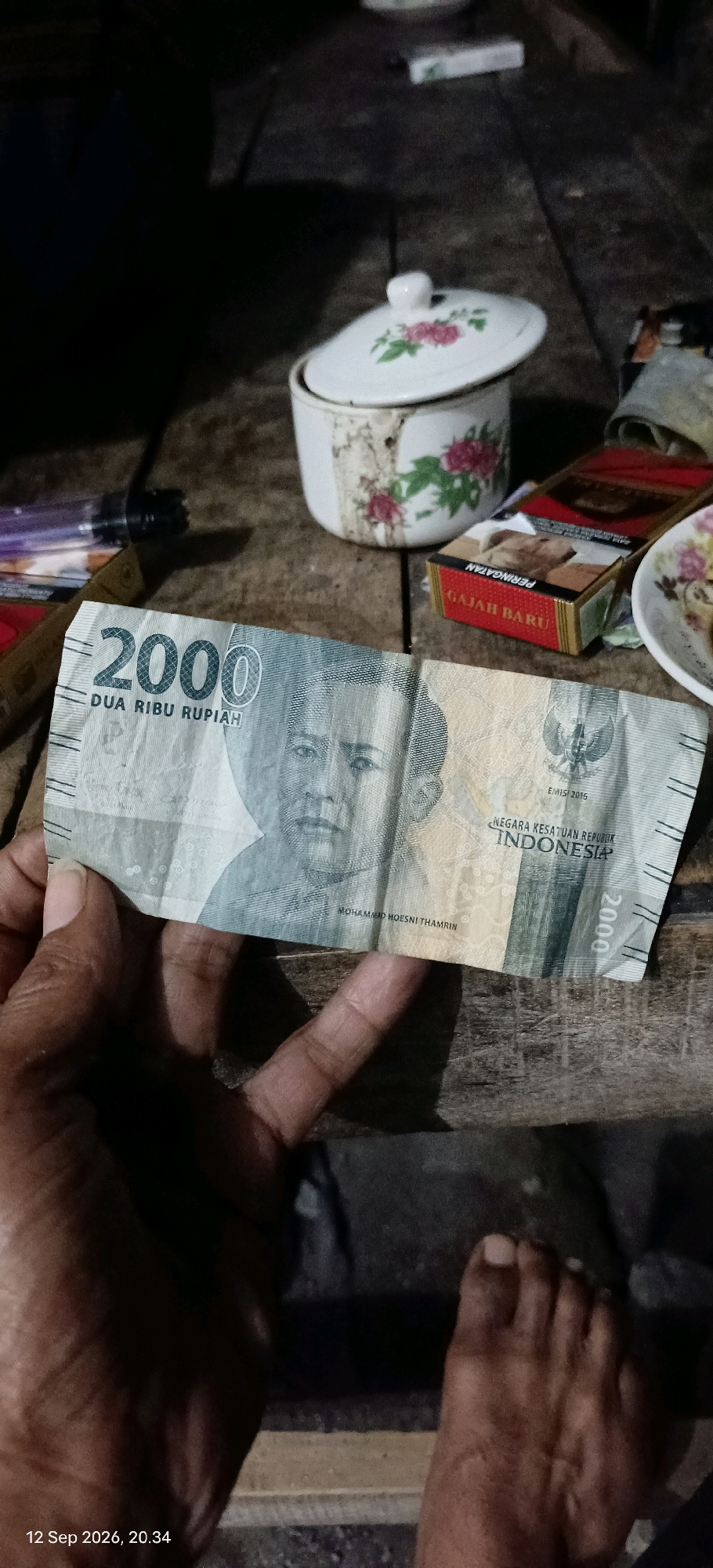 The image depicts a close-up view of a person's hand holding an Indonesian 2000 Rupiah banknote. The banknote features the portrait of Mohammad Hoesni Thamrin and national emblems. In the background, a wooden surface is visible, on which a white ceramic pot with floral decorations and a pack of cigarettes are placed. The lighting suggests an indoor or dimly lit outdoor setting. A date stamp in the lower left corner indicates "12 Sep 2026, 20:34". The overall scene suggests a casual or everyday moment.