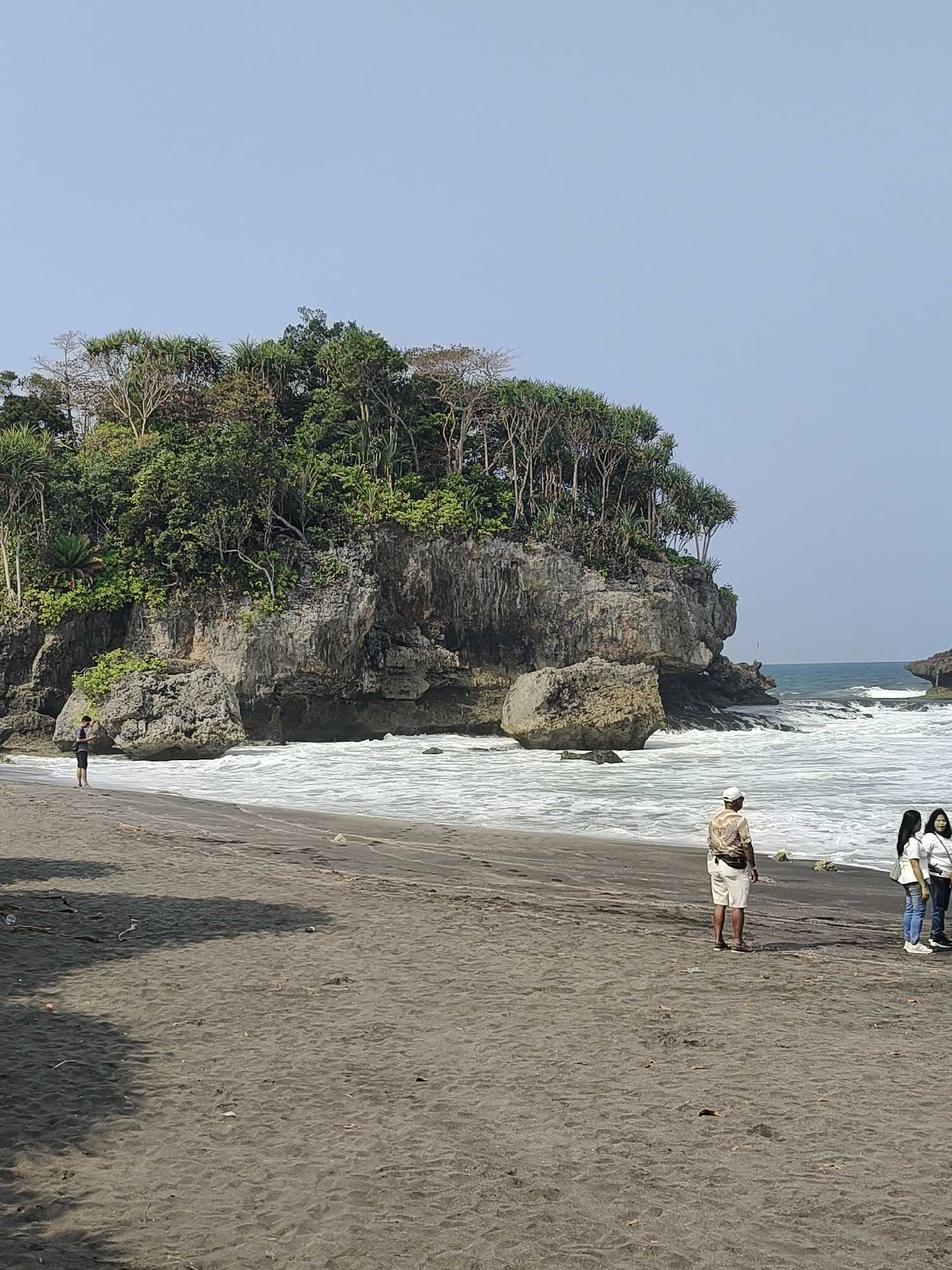 A vertical, eye-level, full outdoor shot captures a beach scene with a prominent rocky outcrop covered in lush green vegetation. The beach itself is composed of dark, coarse sand, with the ocean waves crashing onto the shore in the midground. In the foreground, the dark sand is textured with small debris and shadows cast from an unseen object to the left.
Several individuals are present on the beach. On the left side, a person stands facing away from the camera, near the water's edge, appearing to be looking out at the sea. In the mid-right of the frame, an adult wearing a light-colored hat, a patterned shirt, and white shorts stands facing away from the camera. To their right, two young females stand together, also facing away from the camera. One is wearing a white top and jeans, and the other a white top and dark pants.
The rocky outcrop is the dominant geological feature, with sheer rock faces and boulders at its base. Trees with dense foliage grow from the top and sides of the outcrop. The ocean appears active with whitecaps and swells. The sky is clear and a pale blue. The overall lighting suggests a bright, sunny day. The location is identified as Unknown, Indonesia.
