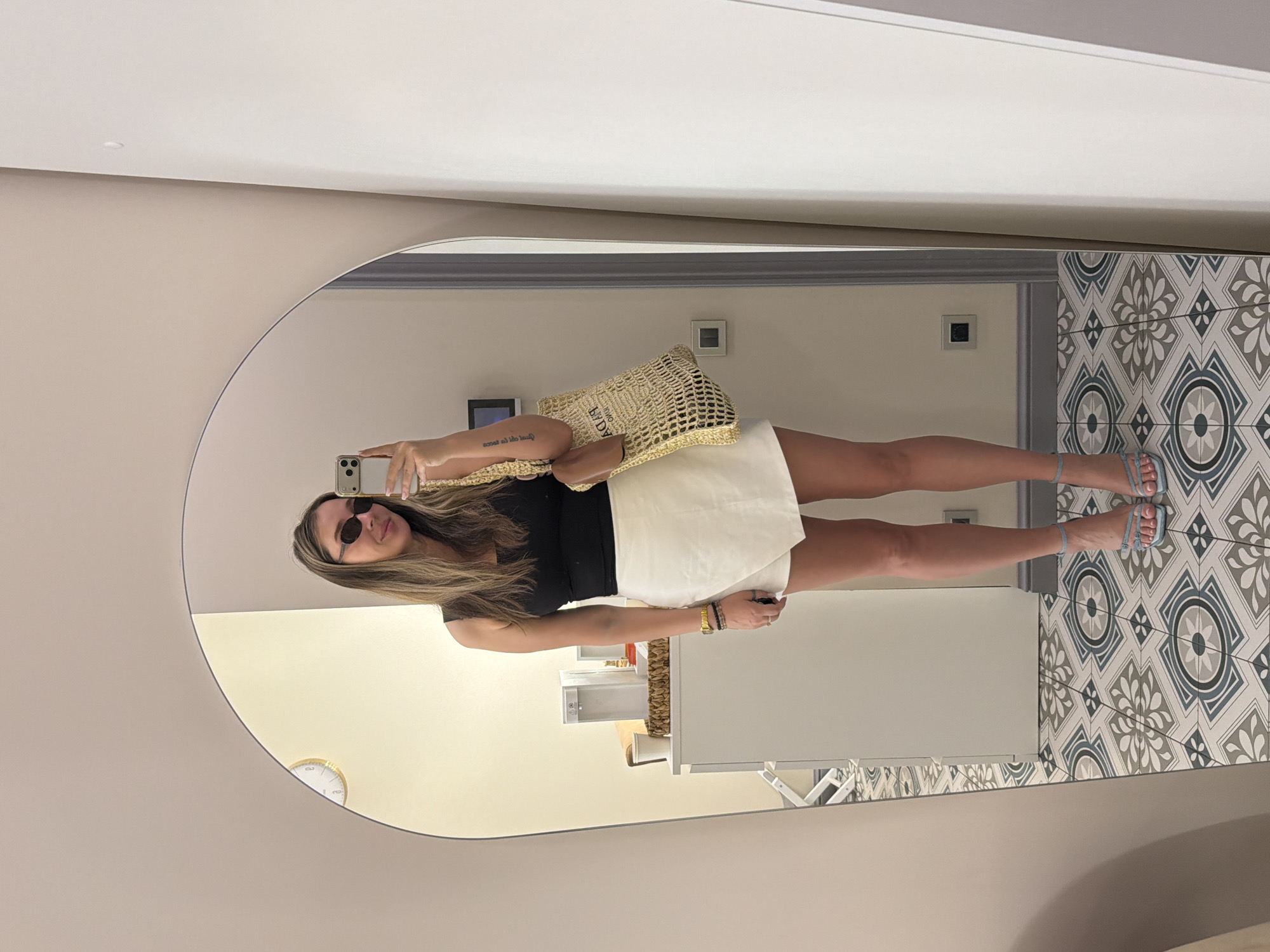 A woman is taking a selfie in a mirror. She is wearing a black top, a white skirt, and blue sandals. She has long blonde hair and is wearing sunglasses. She is holding a straw bag. The background is a tiled wall and a white cabinet. A clock is visible on the wall to the left. The scene appears to be indoors, possibly a bathroom or dressing room. The lighting is bright, suggesting it is daytime. The woman's expression is neutral, and she seems to be posing for the photo. There is no visible text in the image. The location is identified as Moscow, Russia.