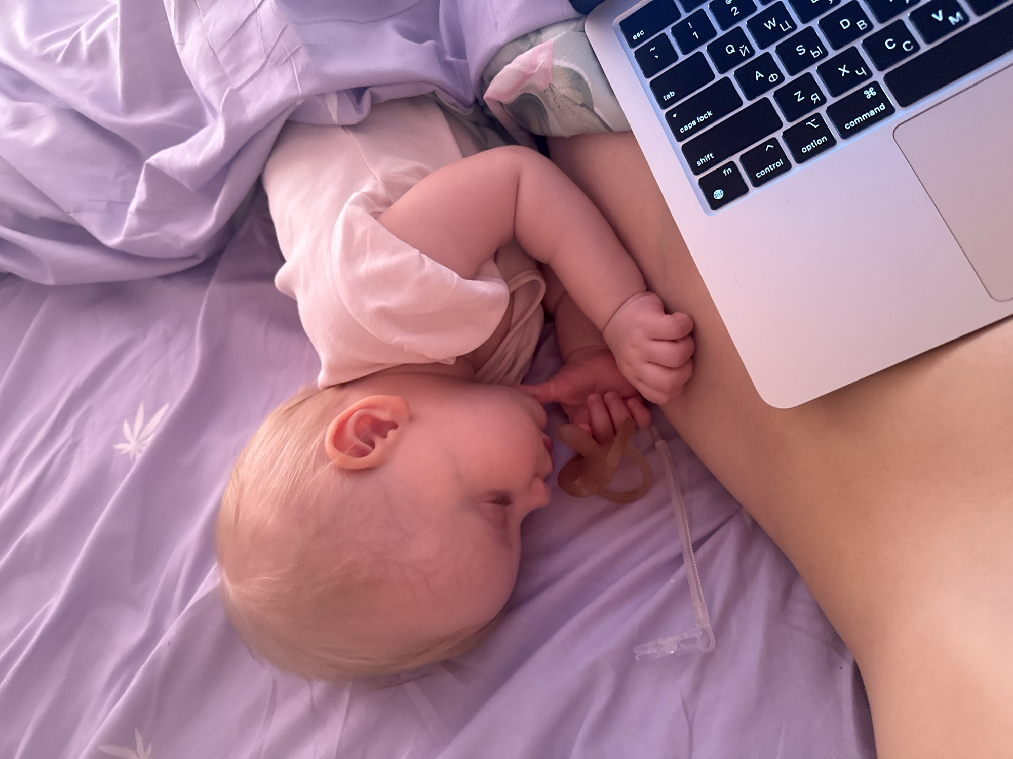 A baby is sleeping on a lavender-colored bed next to a person using a laptop. The baby is on their side, wearing a light-colored onesie and holding a pacifier. The laptop is partially visible, showing its keyboard with Cyrillic letters. The scene appears to be indoors. The exact location in Pokrov, Russia, cannot be determined from the image.