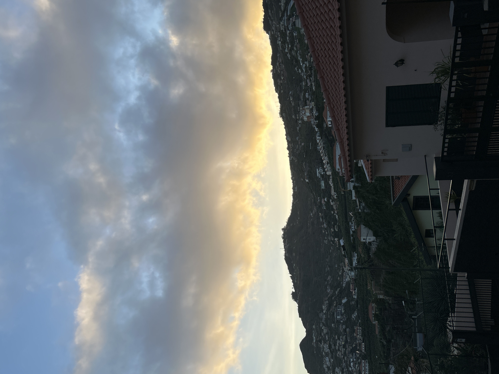 The image shows a view of Câmara de Lobos, Portugal at dusk. The sky is filled with clouds, some of which are illuminated by the setting sun. A mountain is visible in the center of the image, densely populated with buildings. In the foreground, a building with a red-tiled roof and a closed shuttered window is partially visible. A balcony with plants is also visible.