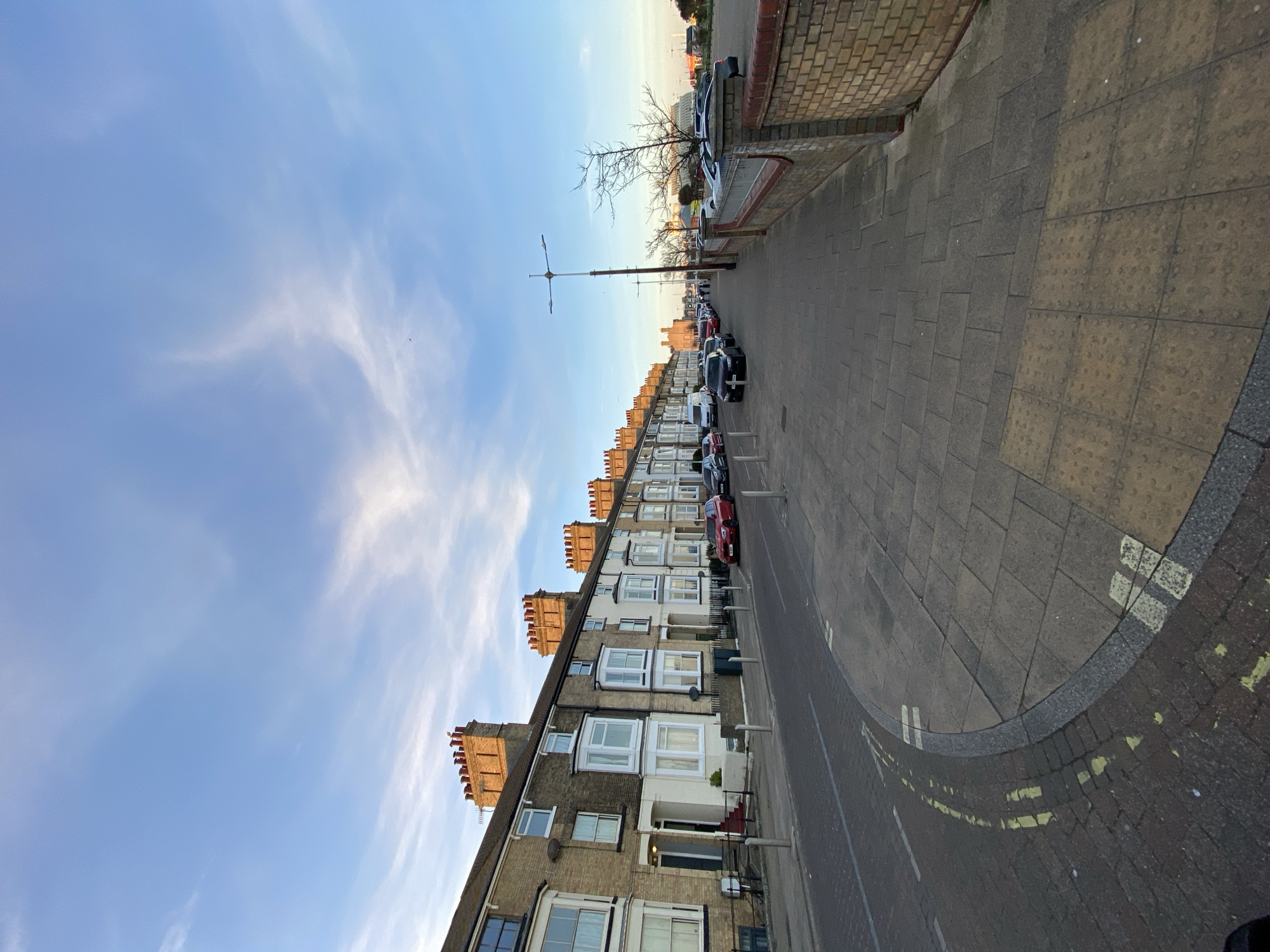 An eye-level, outdoor shot, taken in the town of Felixstowe, in East Suffolk, United Kingdom, displays a residential street. The image is taken from the sidewalk, where the ground is composed of square cobblestones. The sidewalk has white and yellow lines painted onto it. To the left is the view of multiple terraced houses, each with chimneys. Parked along the side of the road are numerous vehicles. In the distance, a bare tree and street lighting are visible. The sky above is mostly blue, with sparse, wispy white clouds. A brick wall runs along the right. No people are visible.