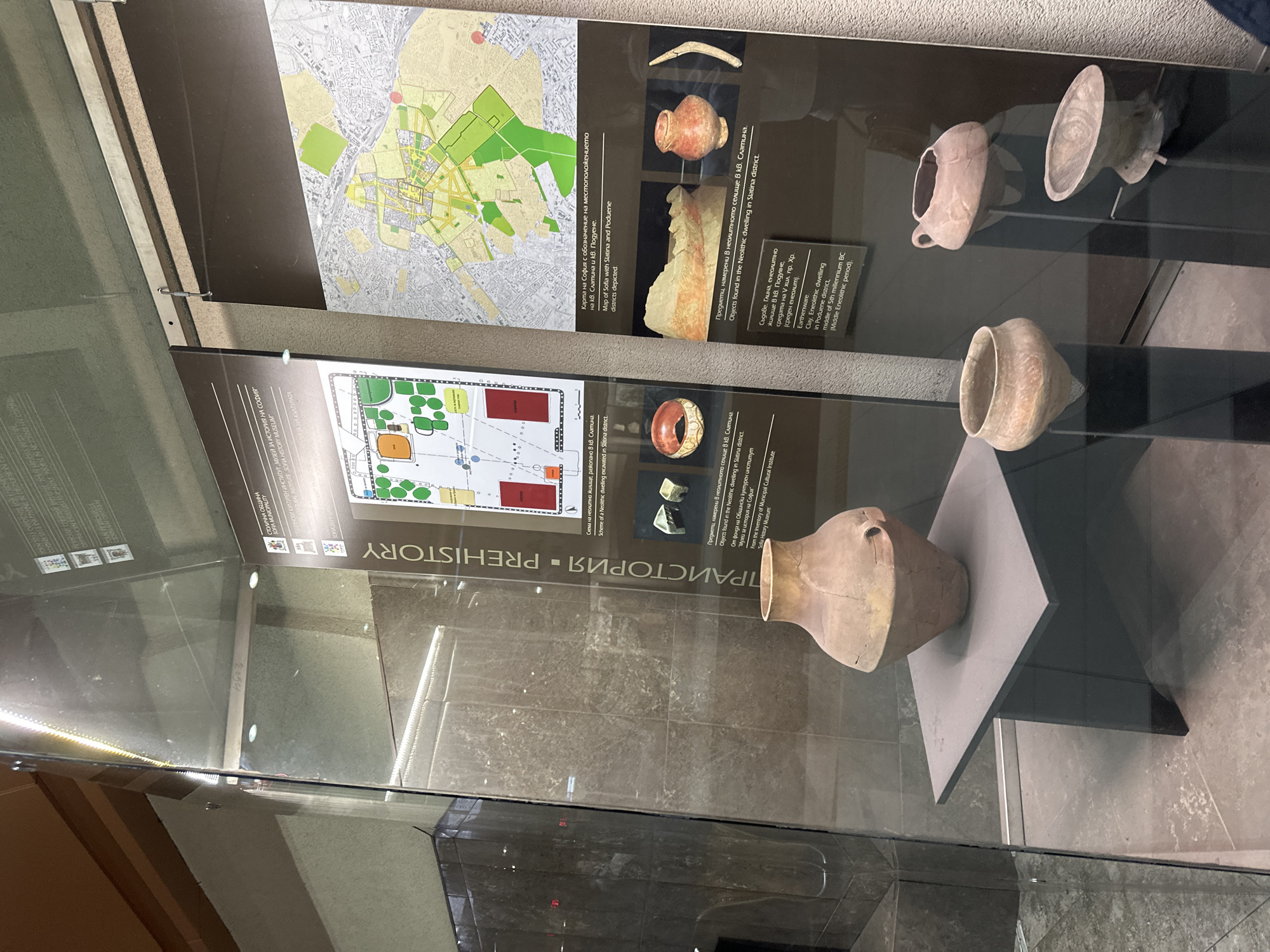 The image depicts a display case in the Sofia History Museum, showcasing artifacts from the Neolithic period found in the Slatina and Poduene districts of Sofia, Bulgaria. The display includes earthenware vessels of various sizes and shapes, along with informational posters containing maps of Sofia indicating the locations of the districts. The posters also contain descriptions of the artifacts and their origins, including details about a Neolithic dwelling excavated in the Slatina district. The artifacts are displayed on stands within the glass case.