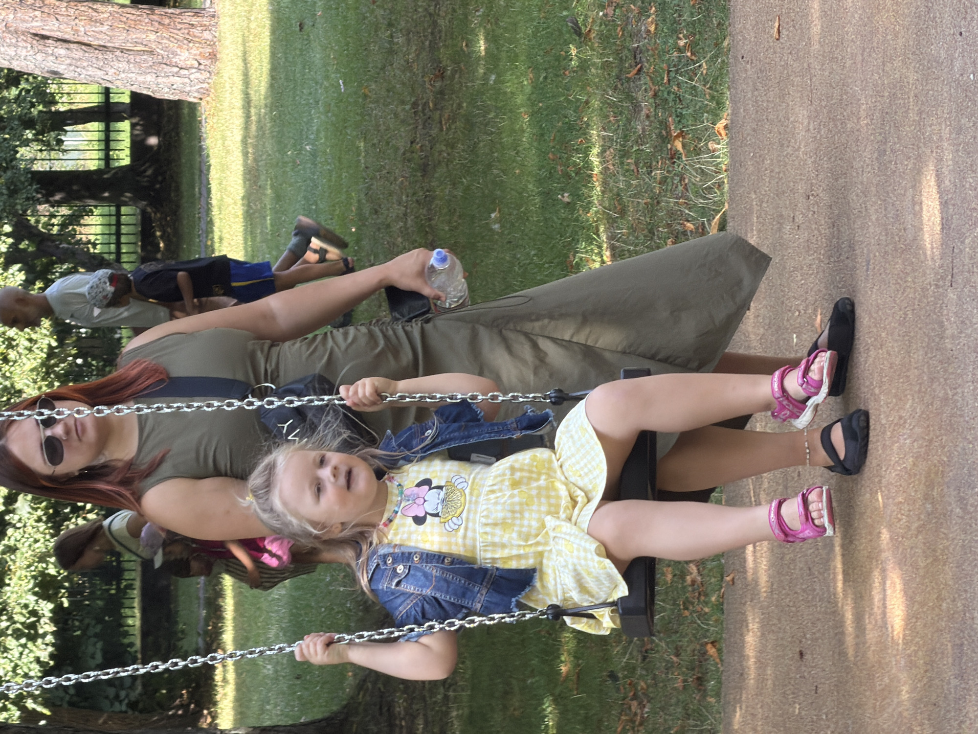 The image shows a group of people in a park-like setting, likely Ventspils, Latvia, based on the provided information. In the foreground, a young girl with blonde hair is sitting on a swing. She is wearing a yellow checkered dress with a Minnie Mouse graphic and a denim jacket. Her feet are dangling, and she is wearing pink sandals. To her left, another person's legs are visible, wearing black shoes. Behind the girl, a woman with red hair and sunglasses is holding a plastic water bottle. She is wearing a long, olive-green dress. In the background, other individuals are blurred, including a man with a shaved head and a child in a black shirt and blue shorts. The environment consists of green grass, trees, and a paved pathway. The scene appears to be a casual daytime outing in a park.