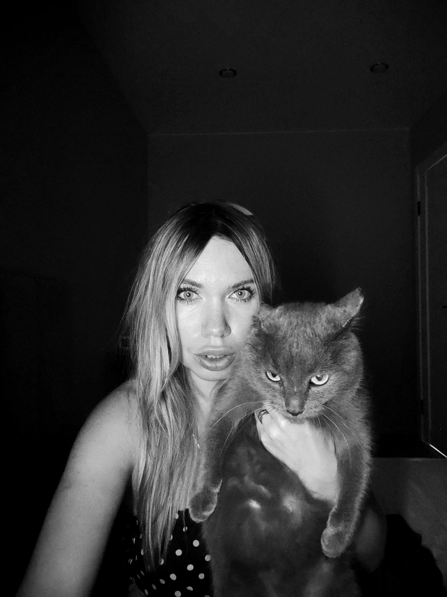 A monochrome, medium shot depicts a fair-skinned woman holding a grey cat. The woman has long blonde hair, light eyes, and is wearing a polka-dot top. Her expression is neutral, with her mouth slightly open. The cat is positioned in her arms, facing the camera, with a stern, narrowed-eye gaze. The background is dark and undefined, suggesting an indoor location. Two ceiling light fixtures can be seen in the upper background along with a partly visible doorway. The specific location in Votkinsk, Russia, cannot be determined from the visual data.