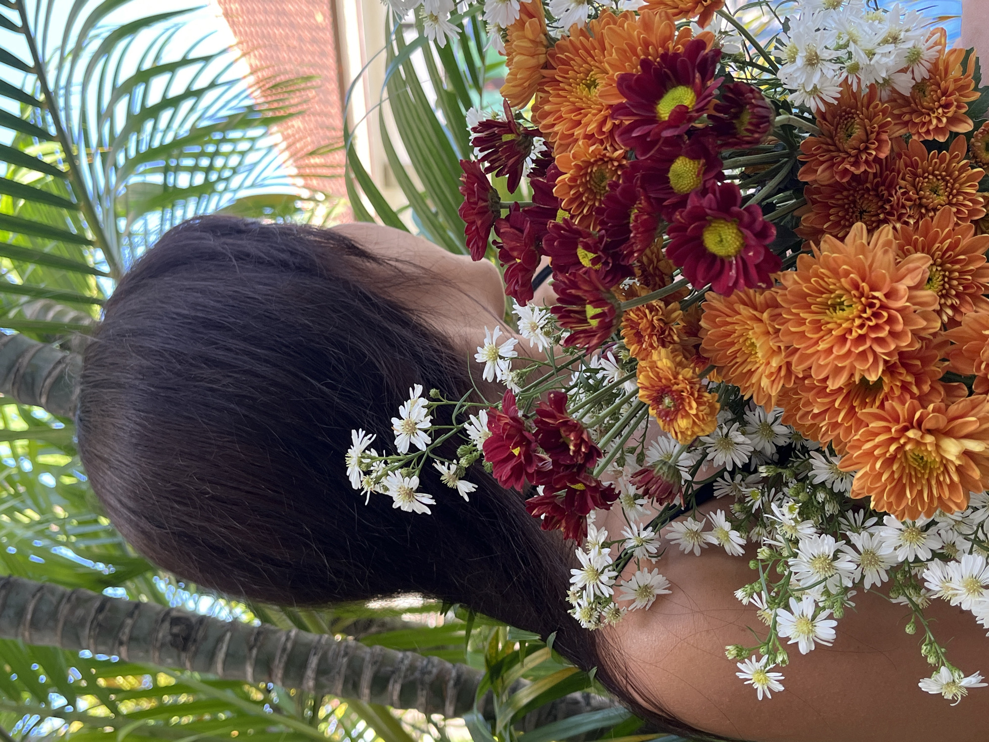 The image displays a close-up portrait of a person's head and shoulder, framed by a vibrant bouquet of flowers in an outdoor, tropical setting. The original image appears rotated 90 degrees counter-clockwise, showing the person's head oriented towards the left.
The main subject is a person with long, dark hair, seen from the back and side. A partial view of their right profile is visible, suggesting they are looking upwards or slightly to the side, surrounded by the flowers.
A large, rich bouquet of flowers is prominently featured, artistically arranged around the person's head and shoulder. The bouquet is dominated by numerous vivid orange and deep burgundy chrysanthemums, characterized by their dense, layered petals. These larger, colorful blooms are interspersed with clusters of smaller, delicate white daisy-like flowers, which add texture and brightness to the arrangement.
The background consists of blurred green palm fronds and other lush foliage, indicating an outdoor, natural environment typical of a tropical region. The scene is brightly illuminated by natural daylight, suggesting clear and sunny weather. The overall impression is one of natural beauty and serenity.
There is no visible text in the media file. The setting is consistent with the provided location context of Kuta Selatan, Indonesia, aligning with a warm and vibrant tropical locale.
