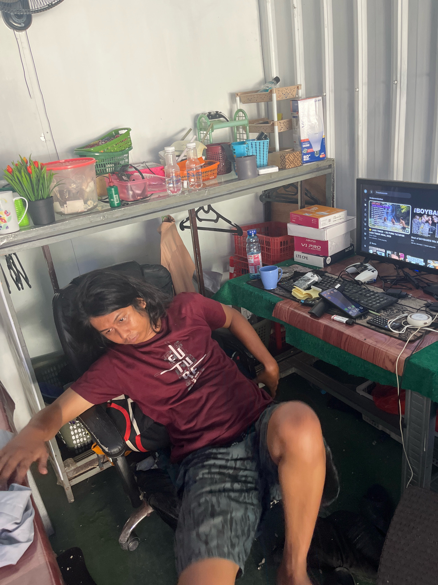 In Sengkang, Indonesia, a person with long, dark hair is seated in a black office chair, wearing a maroon t-shirt with a graphic print and patterned shorts. They are positioned at a desk in what appears to be a cluttered room with various items. On the desk, a computer monitor displays a YouTube video, and there are several boxes, a blue mug, a smartphone, and a keyboard. To the left of the desk, a metal shelf holds multiple baskets, containers, and bottles. The room has a white wall and a corrugated metal wall, suggesting it might be a workshop or a storage area.