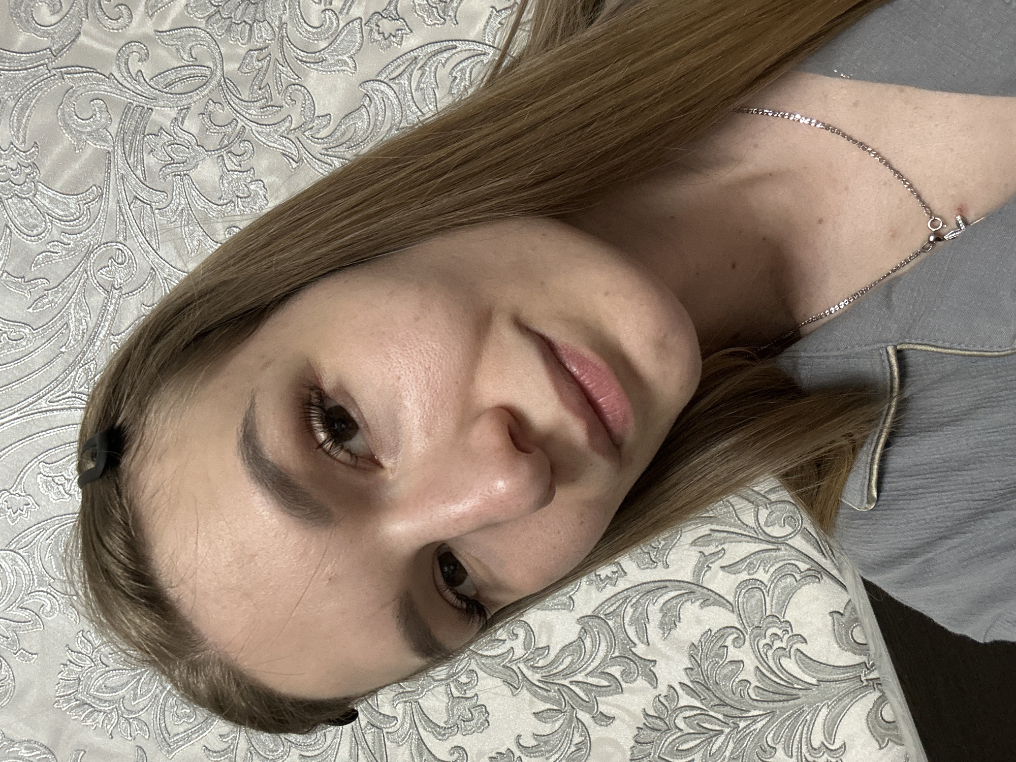 A woman with light brown hair lies on a patterned grey and white bedspread. She wears a grey top and a silver necklace. A black hair clip is visible in her hair. The location is not discernible from the image.
