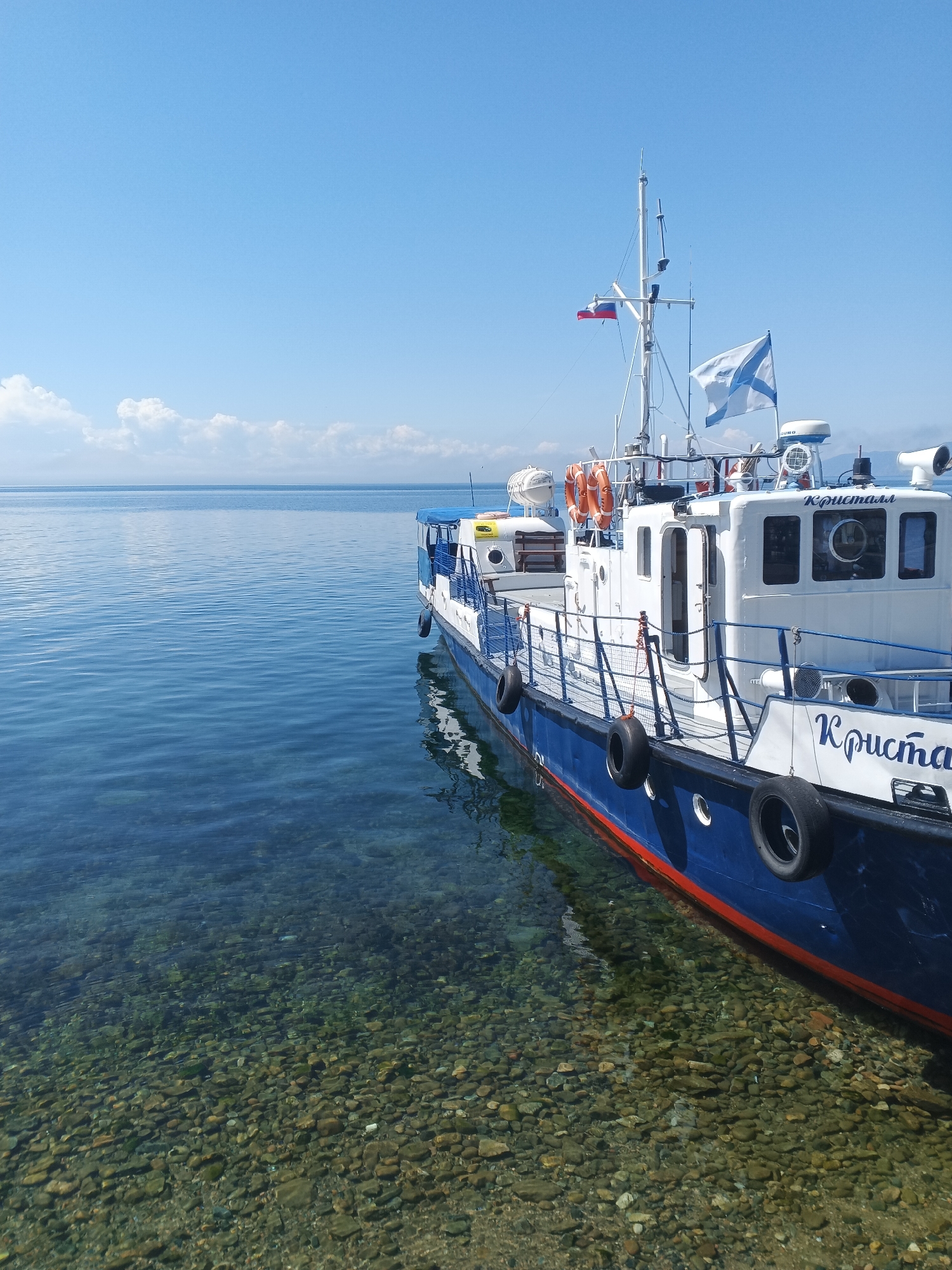 The image depicts a clear, sunny day at the shore of a calm body of water, likely Lake Baikal, given the likely location in Listvyanka, Russia. The foreground reveals the clear, shallow water with a visible, rocky bottom. A blue and white boat with a red waterline is docked near the shore, its hull partially submerged. The boat features life preservers, railings, and windows. The Russian flag and a naval ensign are visible on a mast at the stern of the boat. The background shows a vast expanse of calm, blue water meeting a hazy horizon under a bright blue sky dotted with a few fluffy white clouds. No people are visible in the image.