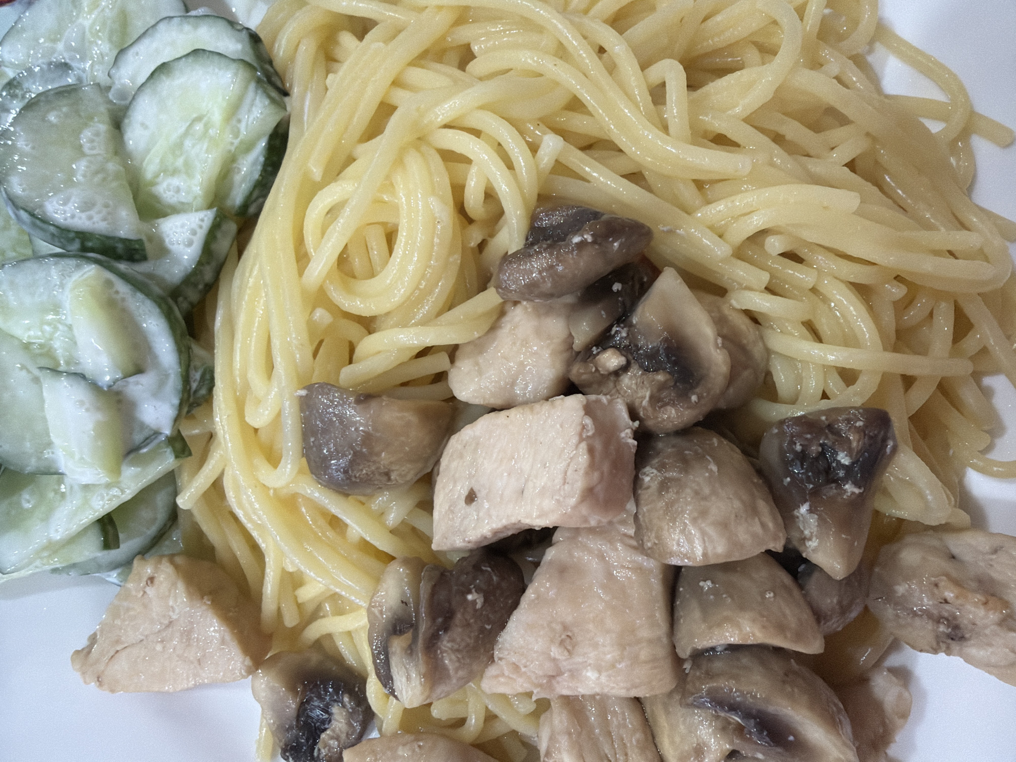 A plate of food is presented in a close-up shot. The plate is filled with spaghetti, cooked chicken pieces, and mushrooms. Beside the spaghetti is a serving of sliced cucumbers with a creamy sauce. The photo appears to be taken in an indoor setting, with the food on a white plate. There is no evidence indicating this photo was taken in Chaikovsky, Russia.