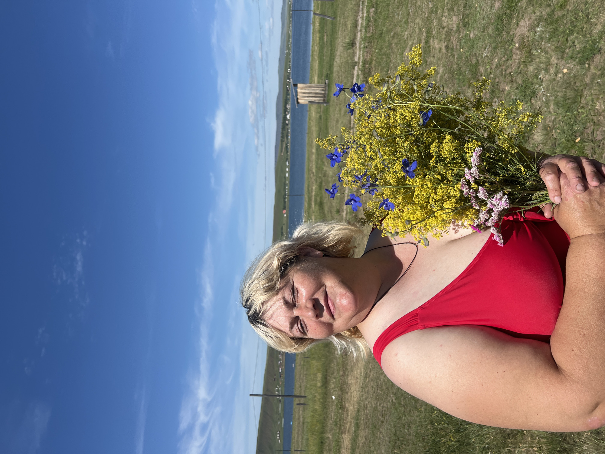 A woman with blonde hair, wearing a red bikini, holds a bouquet of yellow and blue flowers. She is outdoors, with a grassy field and a body of water in the background. The sky is blue with some scattered clouds. The woman is smiling and looking towards the camera. The image appears to be taken on a sunny day. The setting suggests a rural or natural environment.
