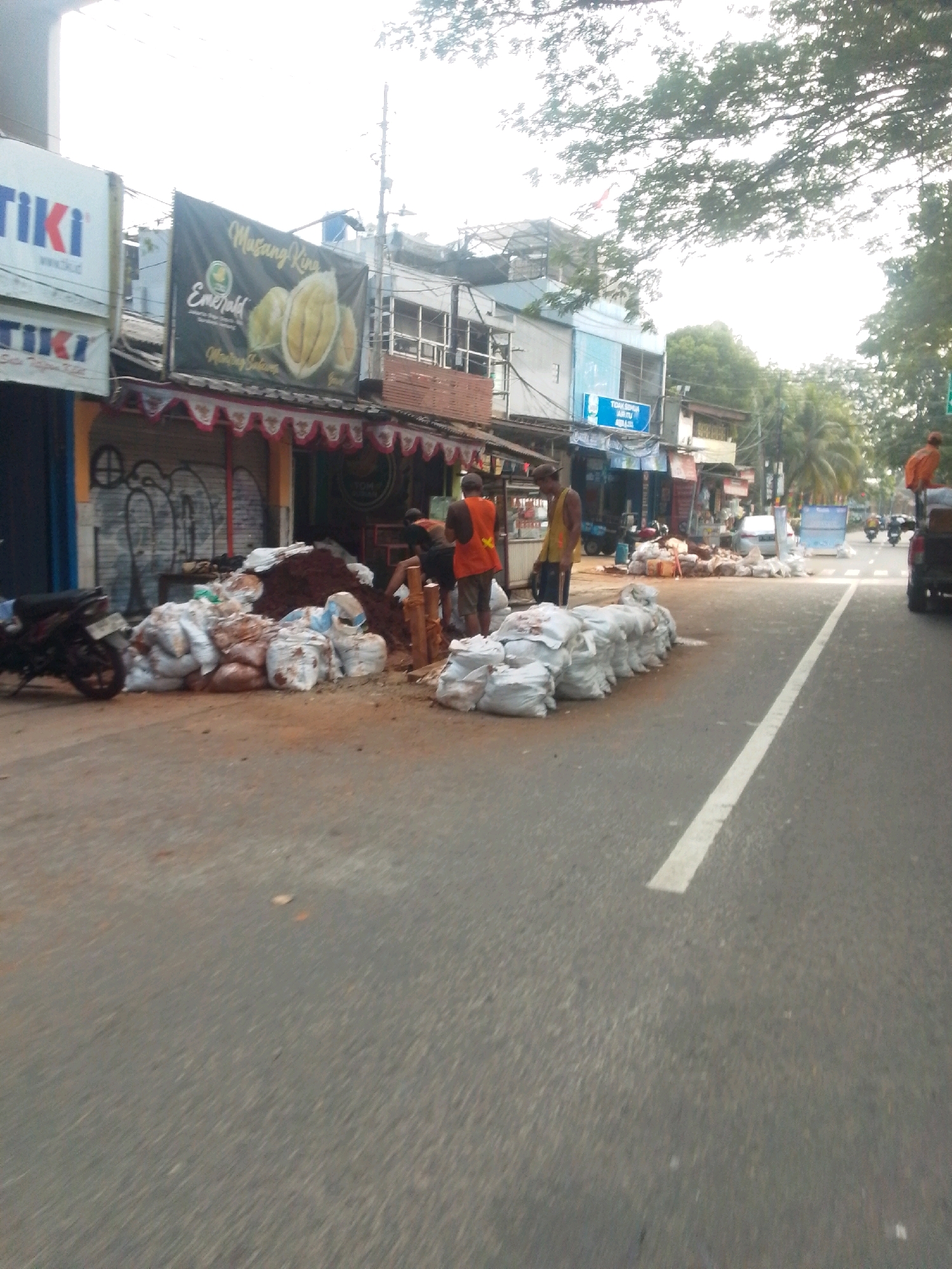 The image depicts a street scene in East Jakarta, Indonesia, with several piles of white sacks filled with soil or sand on the side of the road. A motorcycle is parked to the left of the sacks. Three men are visible: one in an orange vest and dark shorts, another in a yellow vest and brown shorts, and a third partially visible behind the sacks. In the background, there are shops with awnings, signage, and some trees. A few vehicles, including cars and motorcycles, are seen on the road further in the distance. The sky is bright, suggesting daytime.