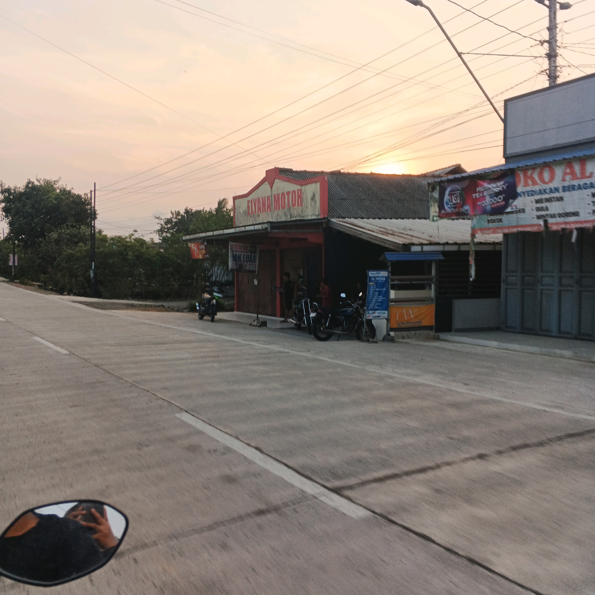 The image depicts a street scene at dusk in Nusajati, Indonesia. The sky is a pale orange, indicative of sunset. On the right side of the street, a shop with the sign "ELYANA MOTOR" is visible, featuring red trim and white lettering. Next to it, another storefront displays a sign reading "KO AL" in large blue letters, with smaller text listing various goods such as "MENISTAK", "MINYAK GORENG", and "ROKOK". Several motorcycles are parked in front of the shops. A narrow road with white lane markings occupies the foreground, and a motorcycle mirror reflects the person taking the photograph. Lush green trees and vegetation line the left side of the road, and utility poles with numerous wires are prominent in the background.
