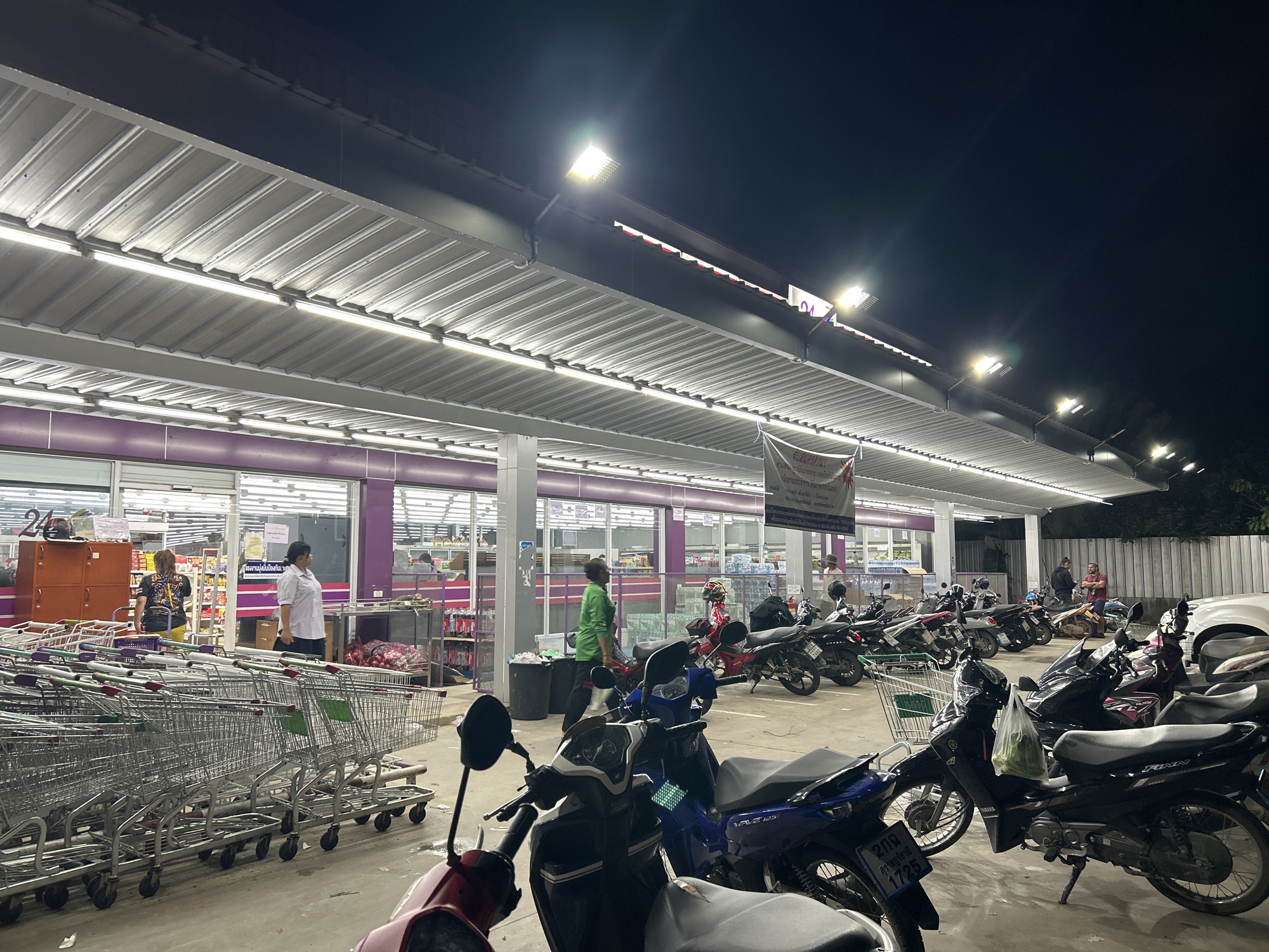 The image shows the exterior of a convenience store in ตำบลเกาะพะงัน, Thailand, at night. Under a well-lit corrugated metal overhang, several people are visible near the storefront, which is visible through large windows. One person is wearing a white shirt, and another a green shirt. Shopping carts are lined up near the store entrance. Numerous motorcycles are parked in front of the store. A banner hangs from the overhang. The sky is dark, indicating it is night.