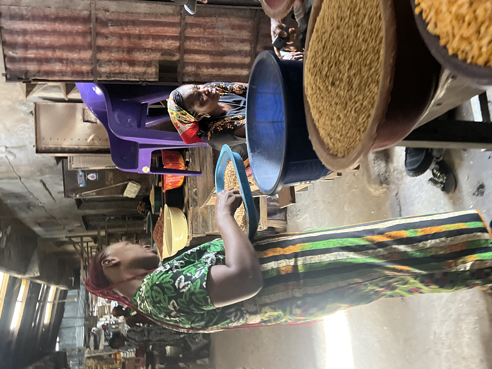 In a bustling market in Nkpor, Nigeria, two women are engaged in the sale of grains. One woman, dressed in a green and black patterned top and a striped skirt, stands facing left, holding a blue bowl filled with grains. The other woman, wearing a headscarf and a dark top with floral patterns, sits on a purple plastic chair behind a blue tub. Both women are positioned near large containers overflowing with grains. The market has a metallic roof and appears busy with other vendors and customers.