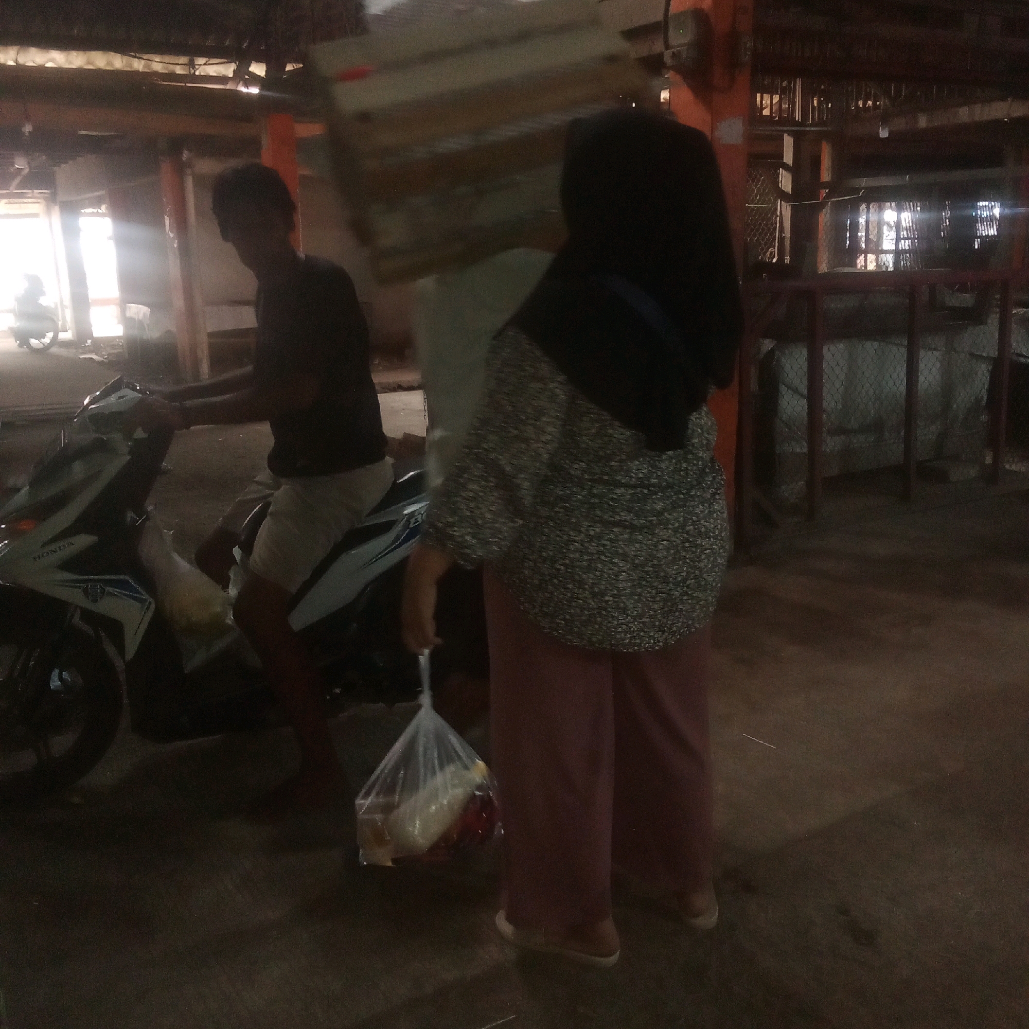 The image depicts a dimly lit outdoor scene, possibly a market or a street in Karawang, Indonesia. In the foreground, a person wearing a black hijab and a patterned shirt is holding a transparent plastic bag filled with food items. They are also wearing loose-fitting pink pants and light-colored slip-on shoes. To the left, a person in a dark t-shirt and light-colored shorts is seated on a white and blue motorcycle. The motorcycle has the "HONDA" logo visible on its side. In the background, there are structures that appear to be market stalls or buildings with wooden beams and corrugated roofing. Some areas in the background are obscured by shadows and low light. The overall impression is of a moment captured in a busy, everyday environment.