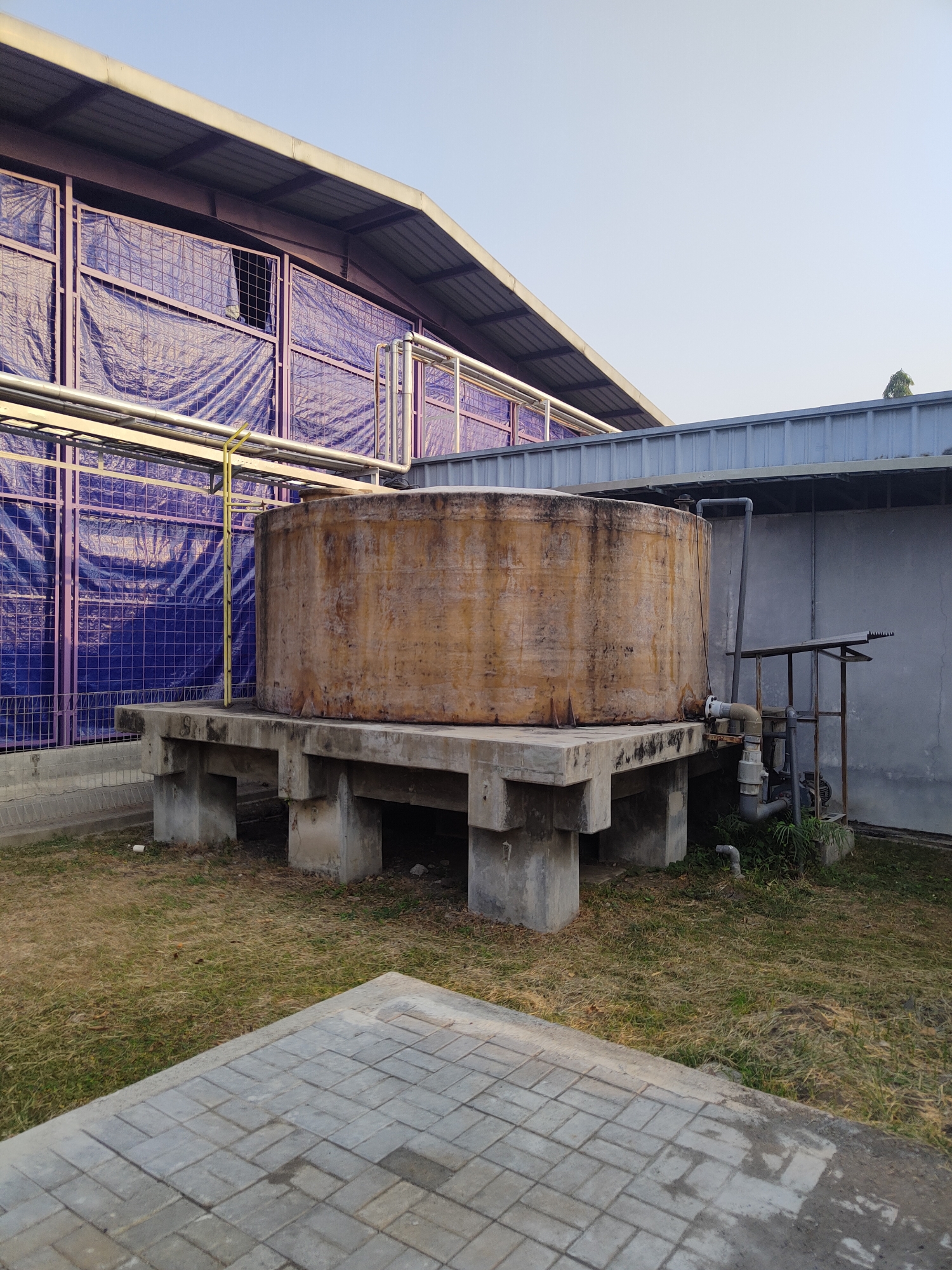 The image shows a large, cylindrical, weathered water tank made of possibly fiberglass or concrete, elevated on a concrete platform supported by multiple concrete pillars. The tank has a rusty or stained appearance. Adjacent to the tank, there is a grey wall and a metal structure with pipes and what appears to be a pump or motor. In the background, a building with a corrugated metal roof is partially covered by a purple tarp and secured with netting. The ground is covered with dry grass and some paving stones are visible in the foreground. The sky is clear and light blue, suggesting it is daytime, possibly late afternoon or early morning due to the angle of the light. The scene appears to be an industrial or agricultural facility located in Raci, Indonesia. There are no people visible in the image.