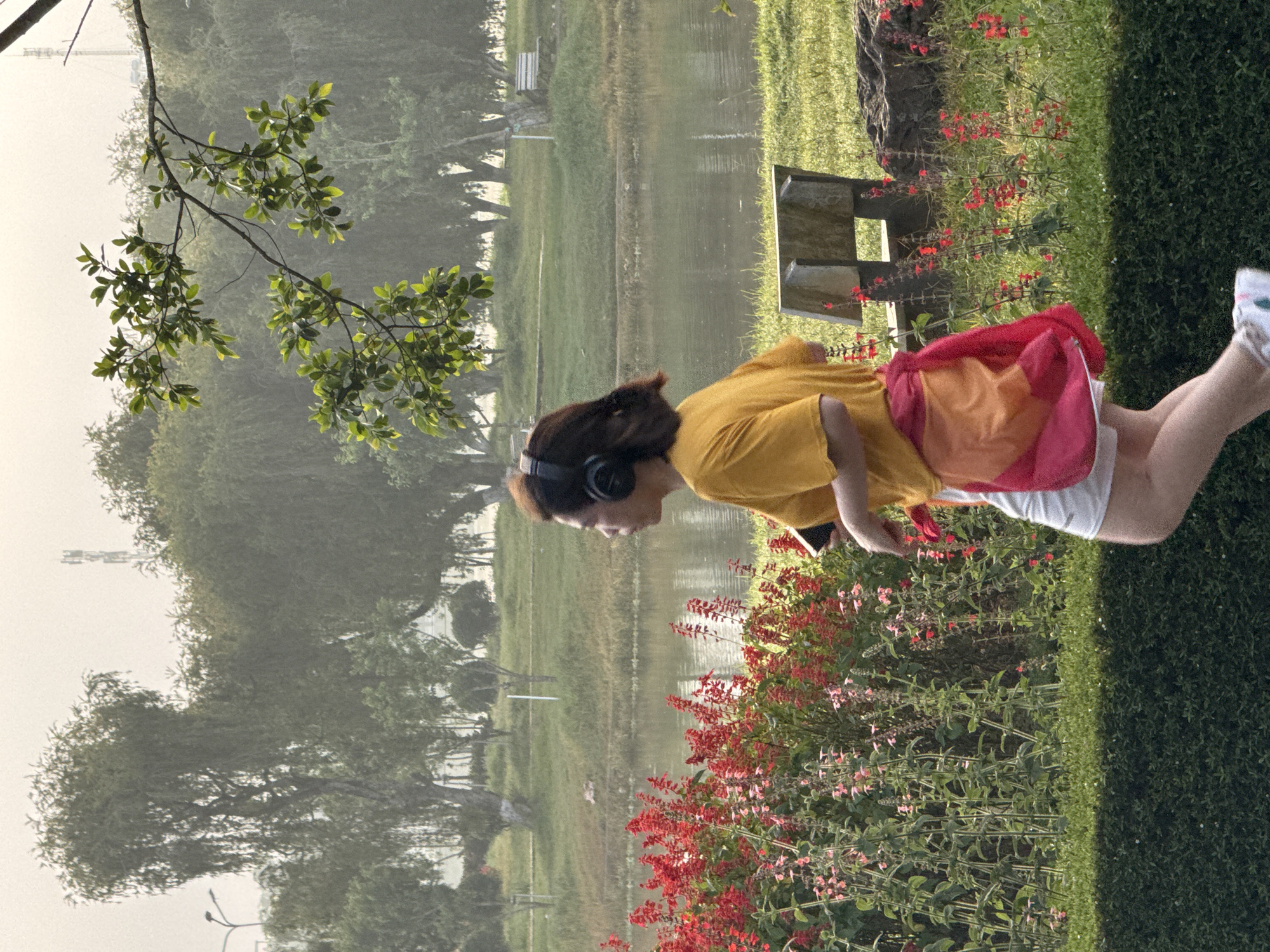 A woman with brown hair, wearing headphones, a yellow t-shirt, and white shorts with a colorful sash tied around her waist, is jogging in a park in Da Lat, Vietnam. She is holding a smartphone in her right hand. The park features a body of water, lush green trees, bushes with red flowers, and a wooden bench. The scene is illuminated by natural light.