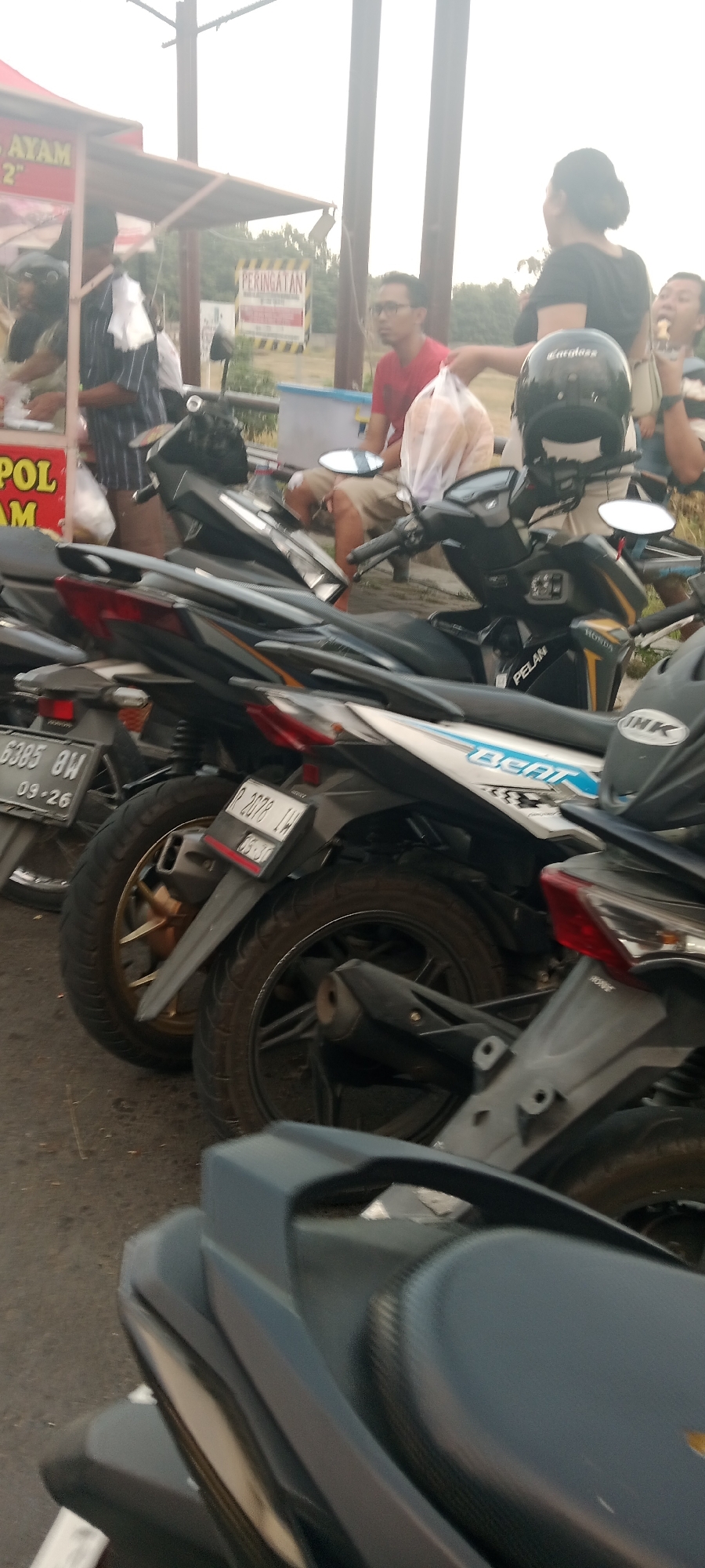 The image shows a street scene in Ngemplak, Indonesia, with several motorcycles parked in front of a food stall. A woman in a black shirt is handing a bag of food to someone off-camera, while a man in a red shirt is observing the transaction. Another person in a blue and white striped shirt is standing near the food stall. A man in tan clothing is bent over near a motorcycle, possibly attending to it. The food stall has a red awning and a sign that says "AYAM". In the background, there is a "PERINGATAN" (warning) sign. The ground is paved and appears to be a road or parking area. The overall atmosphere suggests a casual, everyday scene of commerce and transportation.