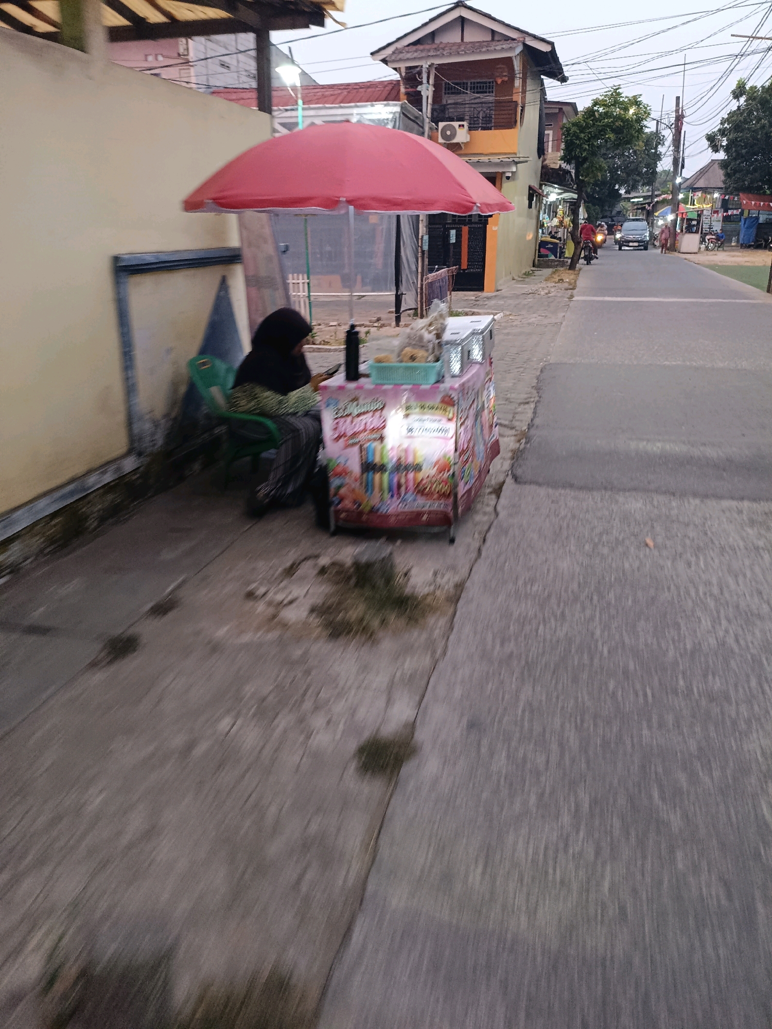 A street vendor is sitting in a green plastic chair under a red umbrella next to her cart in Tangerang, Indonesia. The cart is decorated with colorful images of fruits and candy. A black bottle and some food items are on top of the cart. The vendor is wearing a dark hijab and patterned pants. The street is paved and lined with buildings on the left and right. Several vehicles, including cars and motorcycles, are visible on the street. The sky is overcast. The overall scene suggests a typical street scene in an urban area of Indonesia.