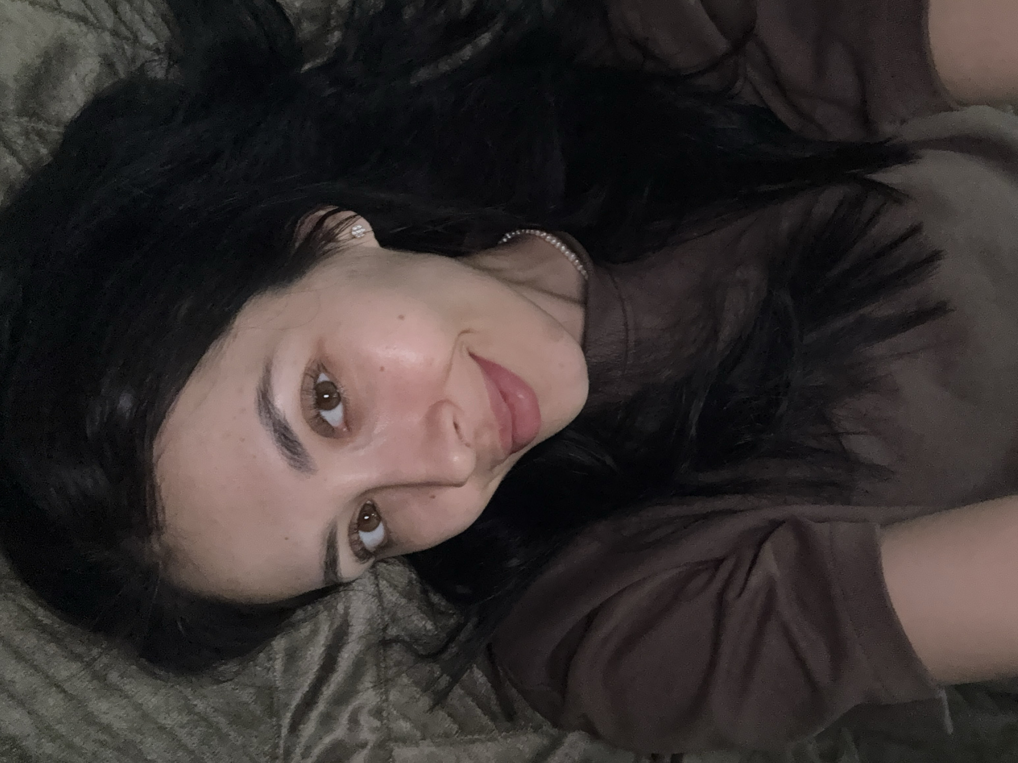A woman with dark hair and brown eyes is lying on a patterned, possibly velvet, surface. She is wearing a brown shirt, a stud earring, and a pearl necklace. She is looking directly at the camera with a slight smile. The image lacks identifying location details to pinpoint Votkinsk, Russia.