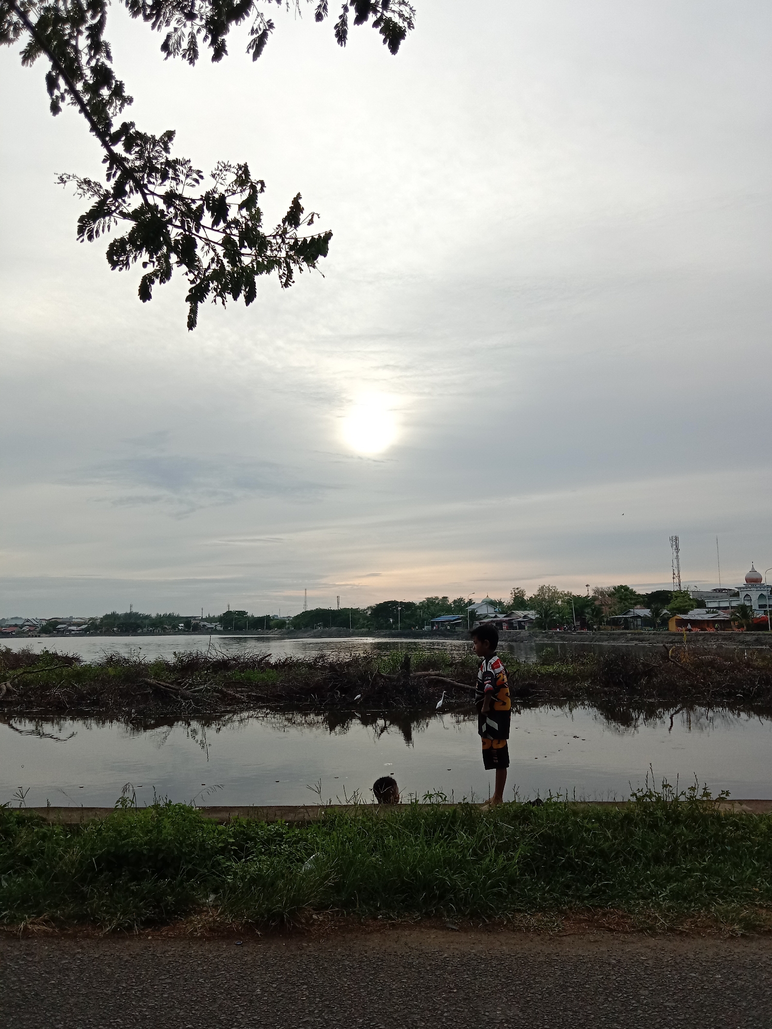 The image shows a waterfront scene in Lhokseumawe, Indonesia. A young boy stands barefoot near the water's edge, wearing a patterned shirt and shorts. The water is lined with debris and vegetation. In the background, buildings, trees, and a mosque dome are visible against a cloudy sky. A tree branch with leaves hangs in the upper left corner of the frame. A paved road and grassy verge are in the foreground.